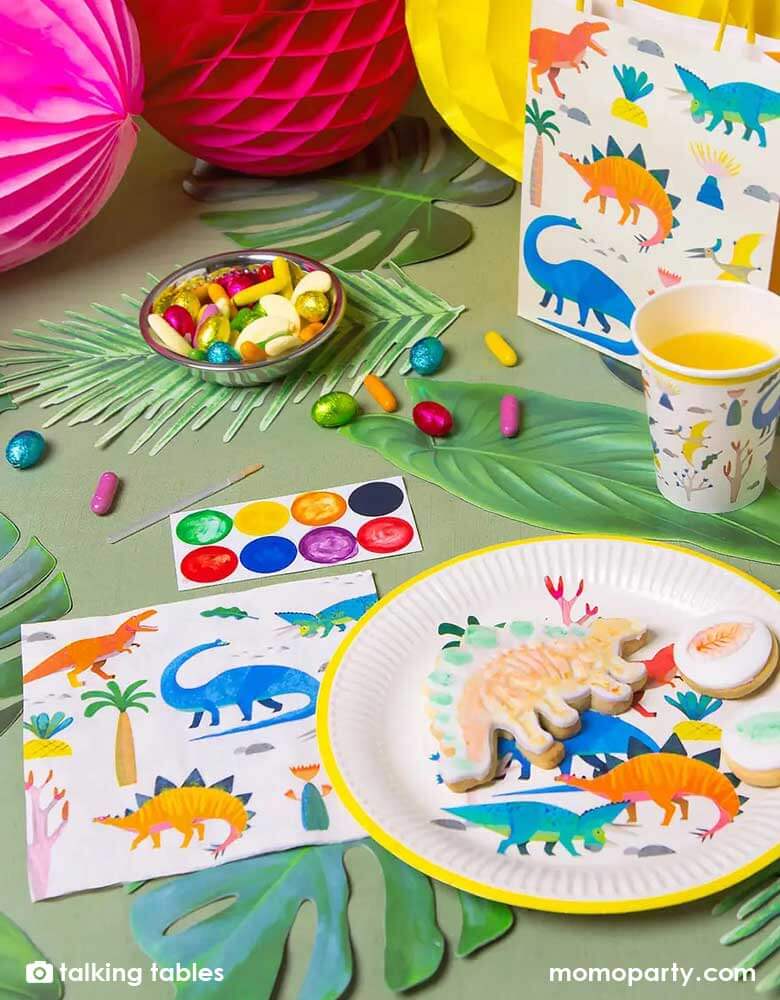 A prehistorical jungle dinosaur themed party table decorated with palm tree leaves and colorful honeycomb balls, this table features Momopartys's 6.5 x 6.5 inches dinosaur party napkins, round dinosaur party plates, cups, and party favor bags by Talking Tables. These dinosaur party supplies feature 5 different jurassic dinosaurs in watercolor style illustrations in bright colors. They're perfect for a kid's dinosaur birthday party.