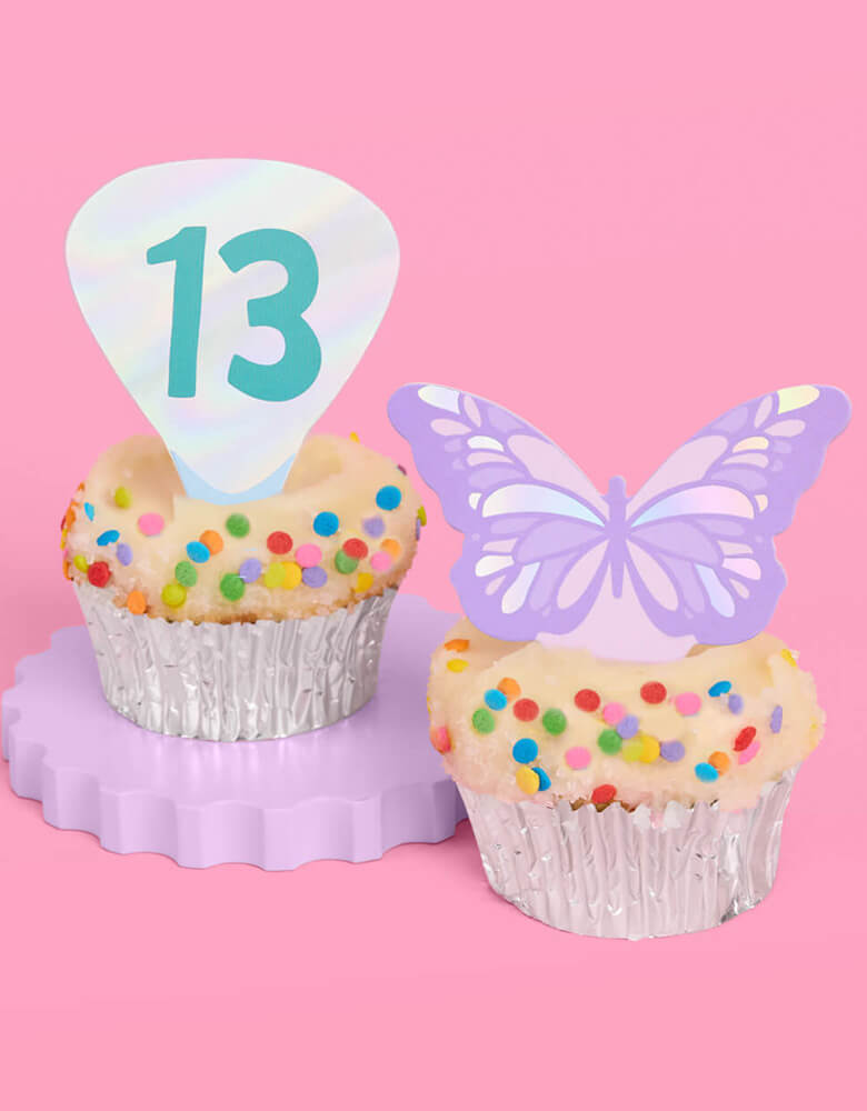 Two birthday cupcakes adorned with Momopartys's Era's Party Cupcake Toppers by Xo, Fetti. Comes in a set of 24, these Taylor Swift-themed dessert/cupcake toppers are with iconic designs including butterflies, red lips, disco balls, cassettes, guitar picks and heart hand symbols. They're perfect for to celebrate your Swiftie girl's special birthday. 
