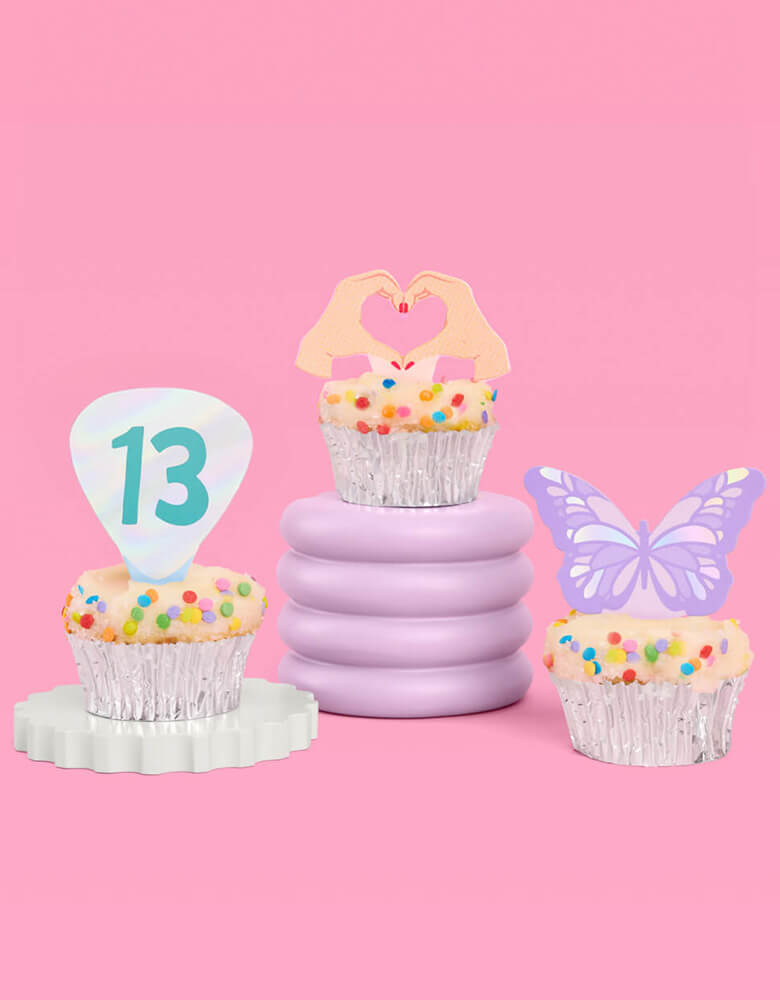 Three birthday cupcakes adorned with Momopartys's Era's Party Cupcake Toppers by Xo, Fetti. Comes in a set of 24, these Taylor Swift-themed dessert/cupcake toppers are with iconic designs including butterflies, red lips, disco balls, cassettes, guitar picks and heart hand symbols. They're perfect for to celebrate your Swiftie girl's special birthday. 