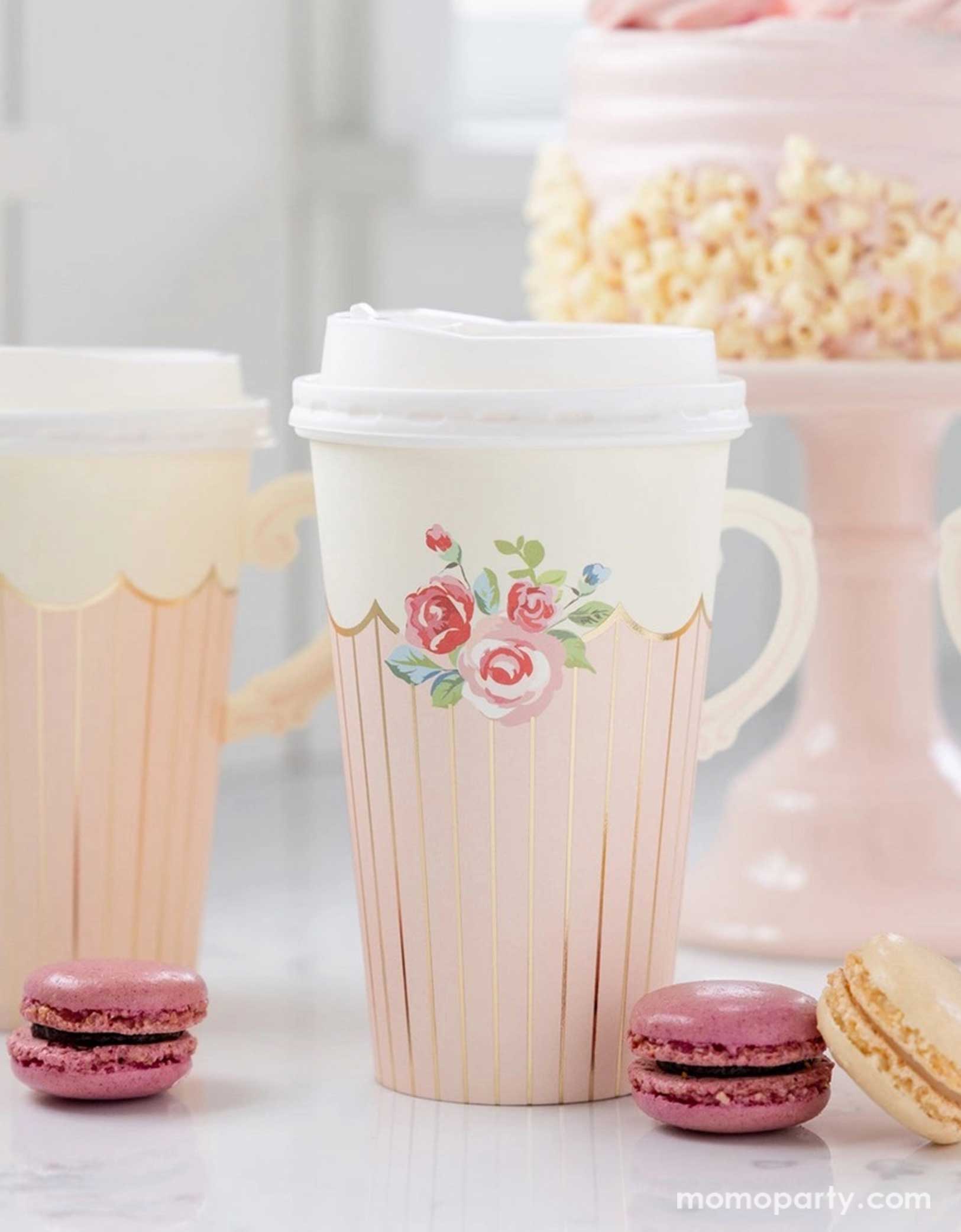 A tea party birthday celebration table set up featuring Momopartys's 12 oz Tea Party Handled Paper Cups by My Mind's Eye. Comes in a set of 8 paper cups, perfect for your tea party on-the-go, these cups feature a clever adhesive handle for easy sipping, with classic rose illustration on the side. Around the paper cups there are some macarons around and a beautiful pale pink cake in the back on a pink cake stand, a perfect setting for a girl's tea party themed birthday celebration. 