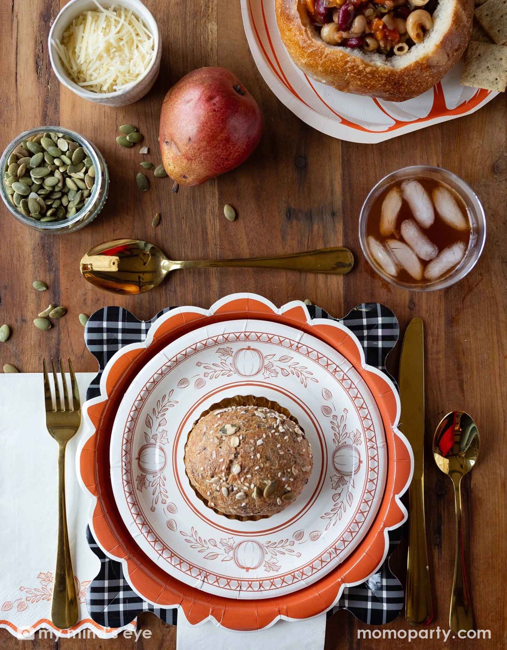 A festive Thanksgiving friendsgiving themed rustic table setting with pumpkin themed plates, bowls, napkins drinks, and gold utensils from Momopartys on a wooden board. With decorative fall themed items including pumpkins, pumpkin seeds, cheese, fall floral and fruit branches, it creates a perfect inspo for a Thanksgiving party, friendsgiving or fall, harvest themed gathering in autumn.