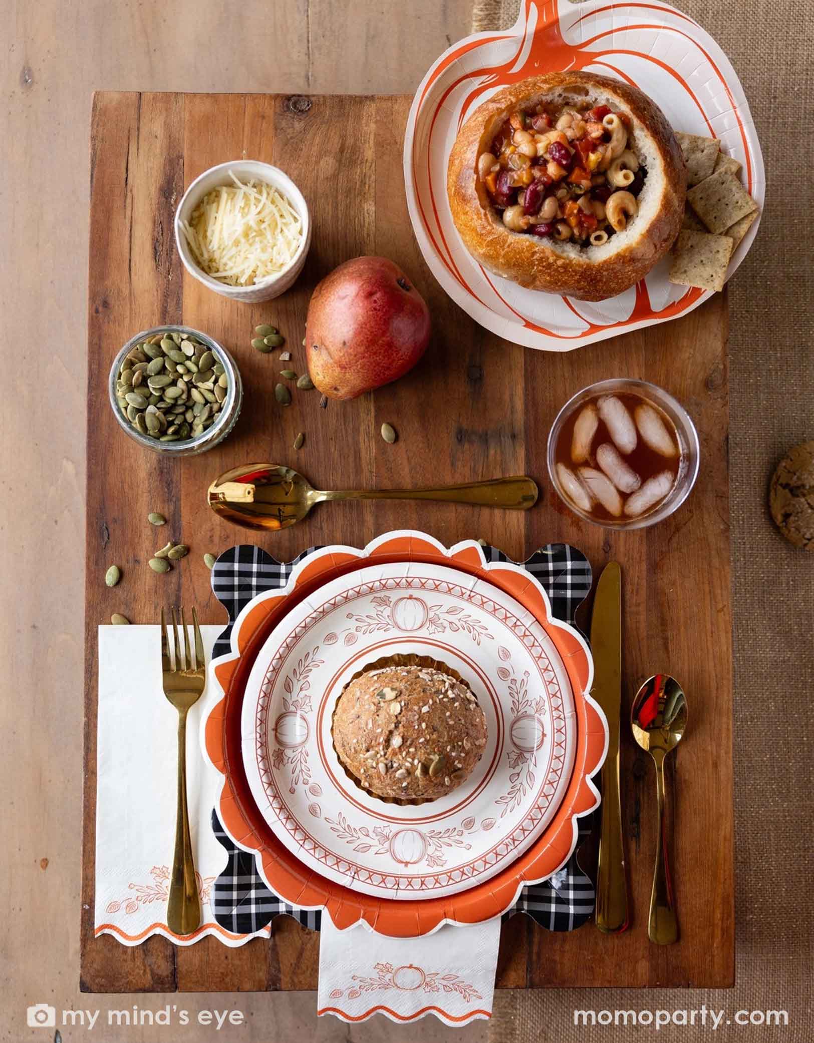 A festive Thanksgiving friendsgiving themed rustic table setting with pumpkin themed plates, bowls, napkins drinks, and gold utensils from Momopartys on a wooden board. With decorative fall themed items including pumpkins, pumpkin seeds, cheese, fall floral and fruit branches, it creates a perfect inspo for a Thanksgiving party, friendsgiving or fall, harvest themed gathering in autumn.