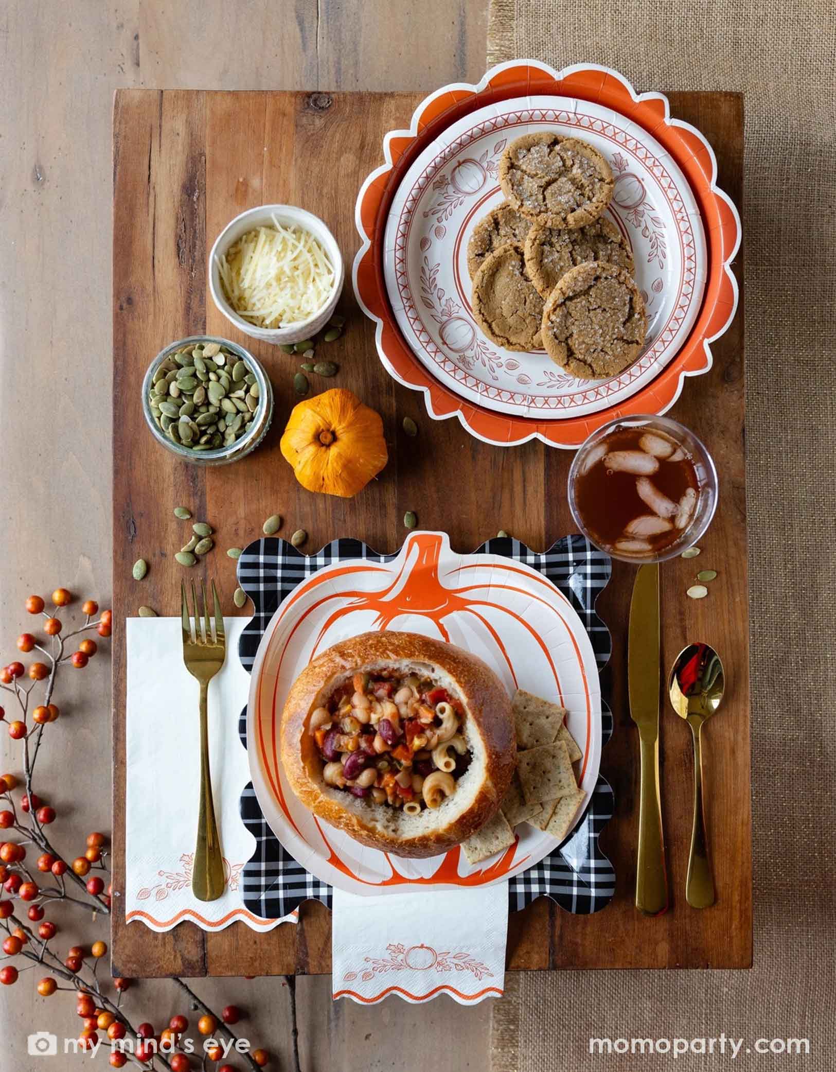 A festive Thanksgiving friendsgiving themed rustic table setting with pumpkin themed plates, bowls, napkins and gold utensils from Momopartys on a wooden board. With decorative fall themed items including pumpkins, pumpkin seeds, cheese, fall floral and fruit branches, it creates a perfect inspo for a Thanksgiving party, friendsgiving or fall, harvest themed gathering in autumn.