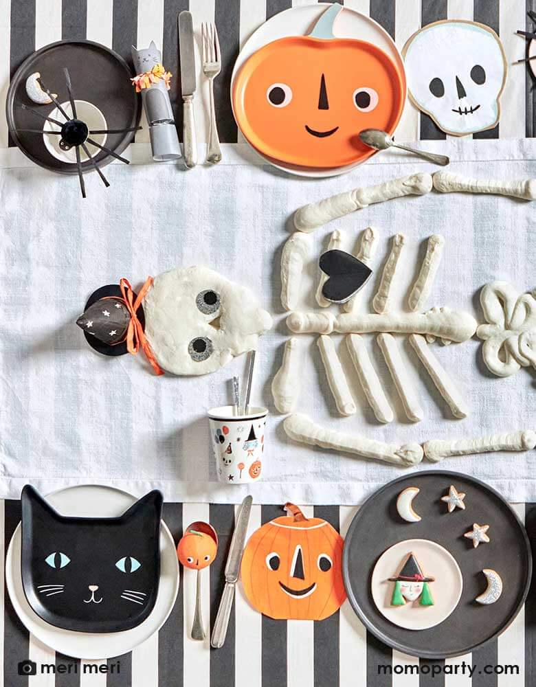 An overhead shot of a Halloween party table featuring Momopartys's modern Halloween tableware including black striped tablecloth, This is Halloween! tableware, napkins, and plates in the classic Halloween icons including pumpkins, skulls, black cats and spiders. In the middle there's skeleton shaped cookies in the middle as the centerpiece making this a modern yet playful tableset for a kid's friendly Halloween table setting ideas.