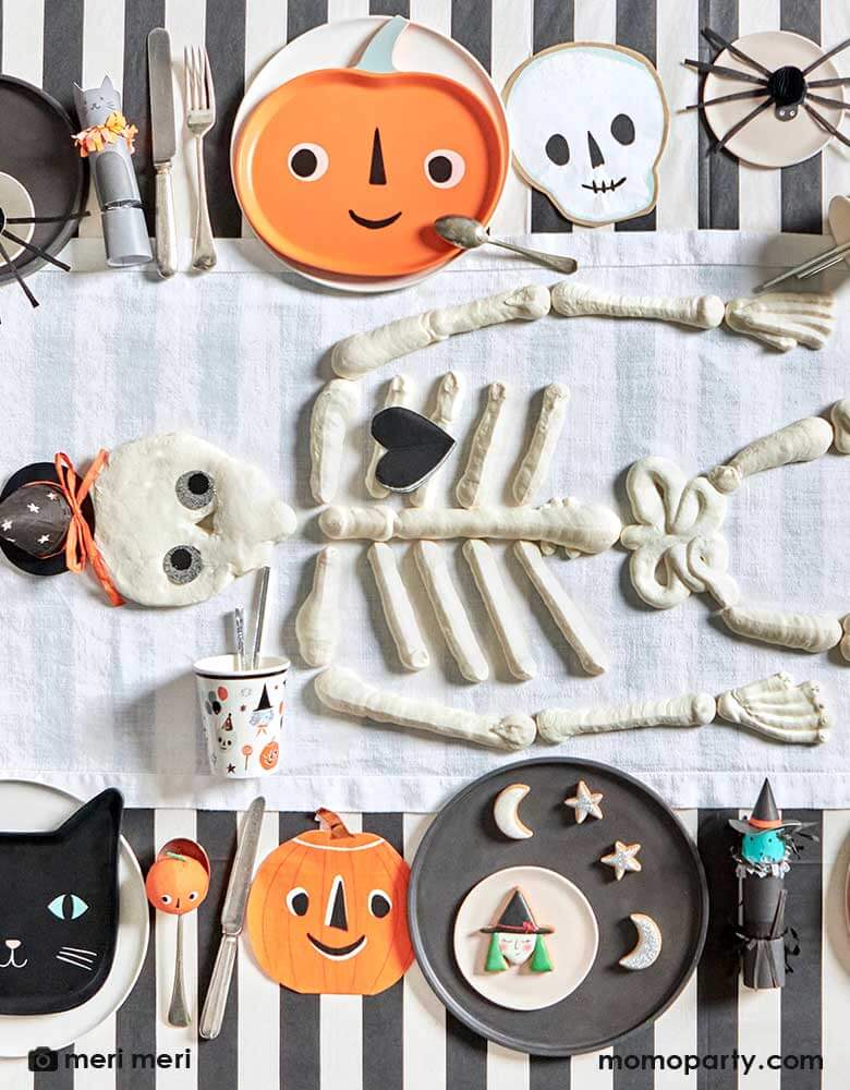  An overhead shot of a Halloween party table featuring Momopartys's modern Halloween tableware including black striped tablecloth, This is Halloween! tableware, napkins, and plates in the classic Halloween icons including pumpkins, skulls, black cats and spiders. In the middle there's skeleton shaped cookies in the middle as the centerpiece making this a modern yet playful tableset for a kid's friendly Halloween table setting ideas.