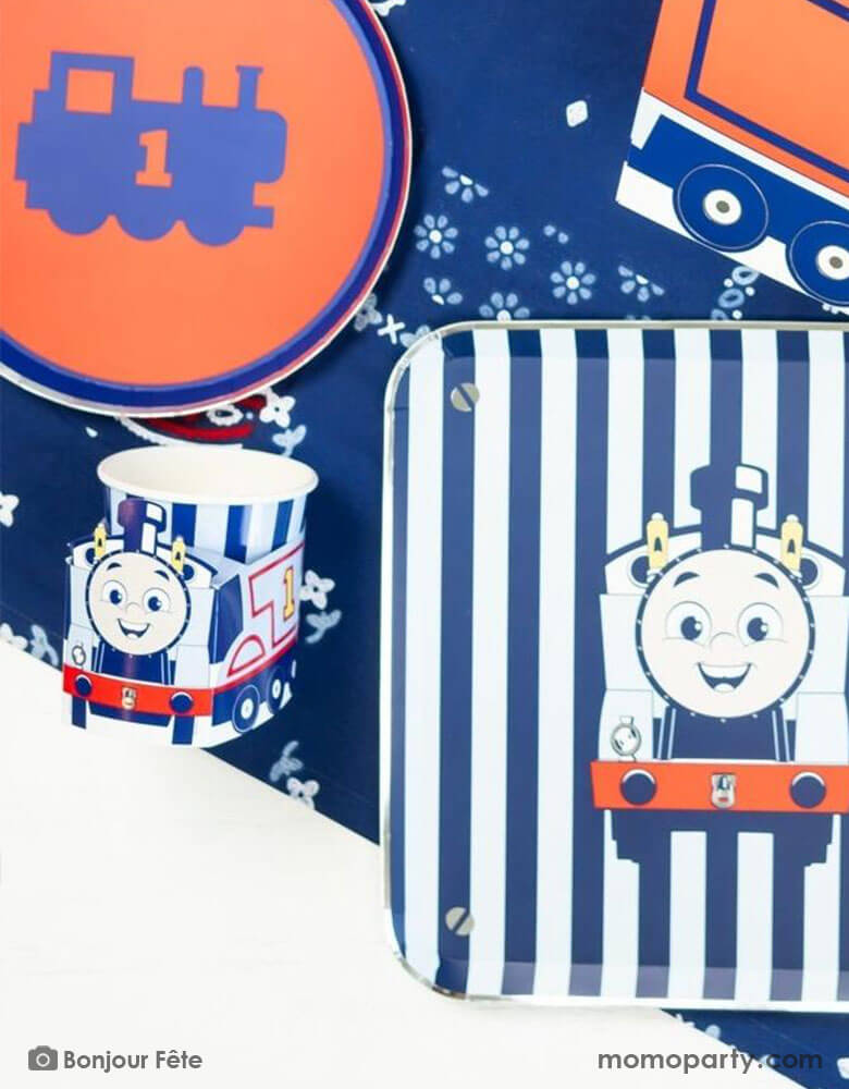 A kid's train themed birthday party table with Momopartys's Thomas and Friends party supplies including the 3D party cups, train railroad round red side plates, Thomas & Friends blue striped dinner plates, and blue floral table runner undernewath. A perfect combination for your toddler's chugga chugga choo choo train themed celebration! All aboard!