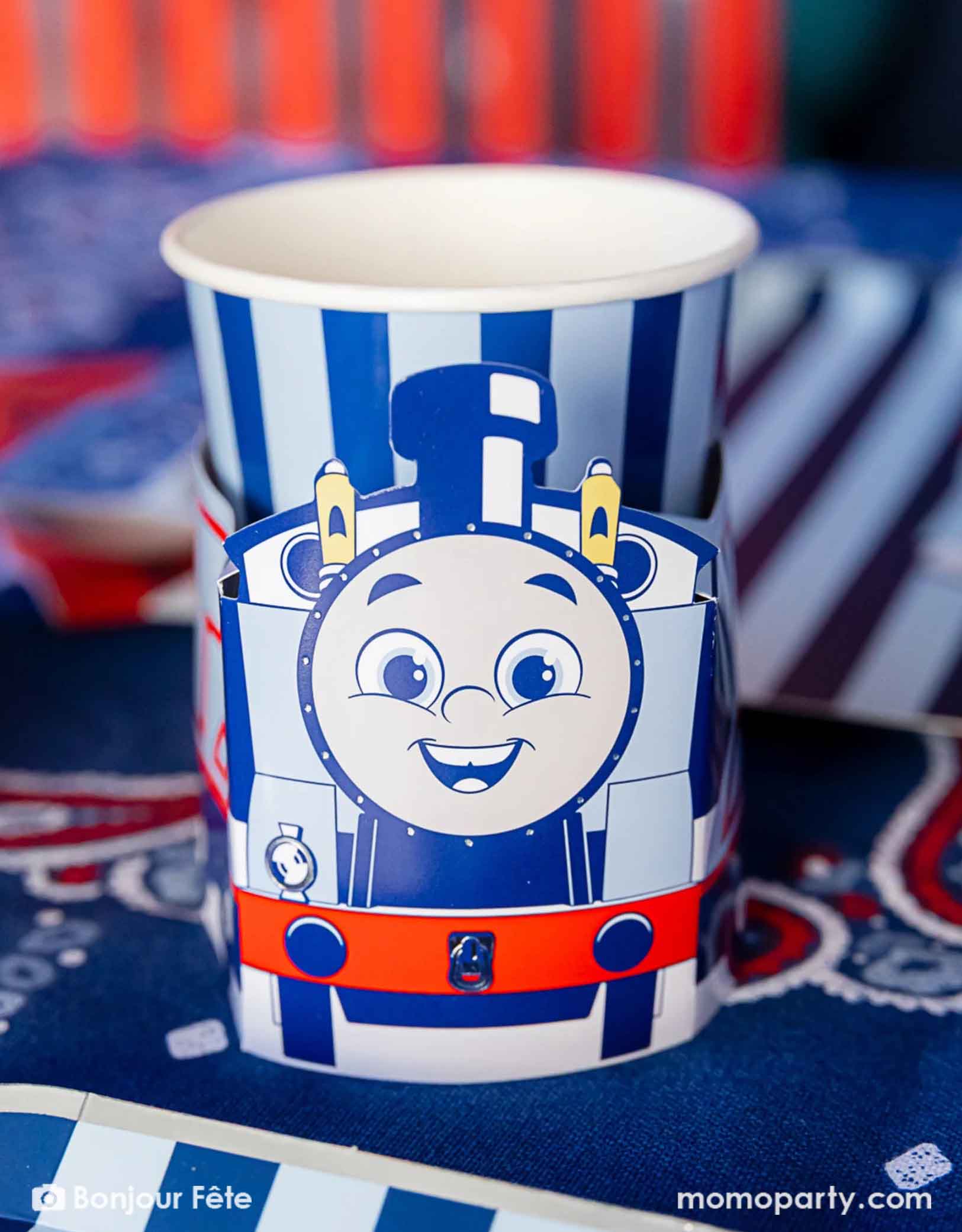 A close-up shot of the Thomas the Train and Friends 3D party cup in a festive Thomas the Train and Friends themed train party. On the table there are various Thomas the Train themed party supplies from Momopartys including party tableware of plates, napkins treat boxes. The party table is covered with bandana inspired tablecloth to match the theme, making this a perfect table setting idea for a toddler's train themed birthday party celebration.