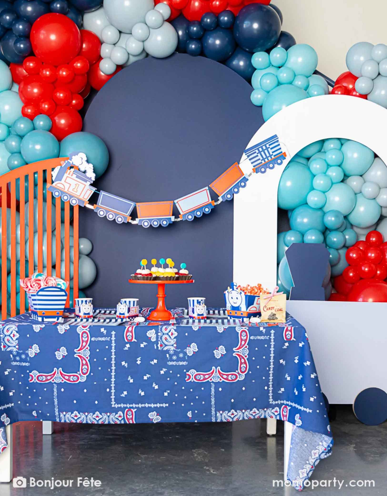 An adorable and festive Thomas the Train and Friends themed train party set up featuring Momopartys's Thomas the Train themed party supplies including party tableware of plates, cups, napkins treat boxes and party garland. The party table is covered with bandana inspired tablecloth to match the theme. In the middle of the table there's a cake stand holding cupcakes in yellow and red colors topped with pom pom toppers. In the back of is a blue & red balloon garland,  a perfect inpo for kid's train party.