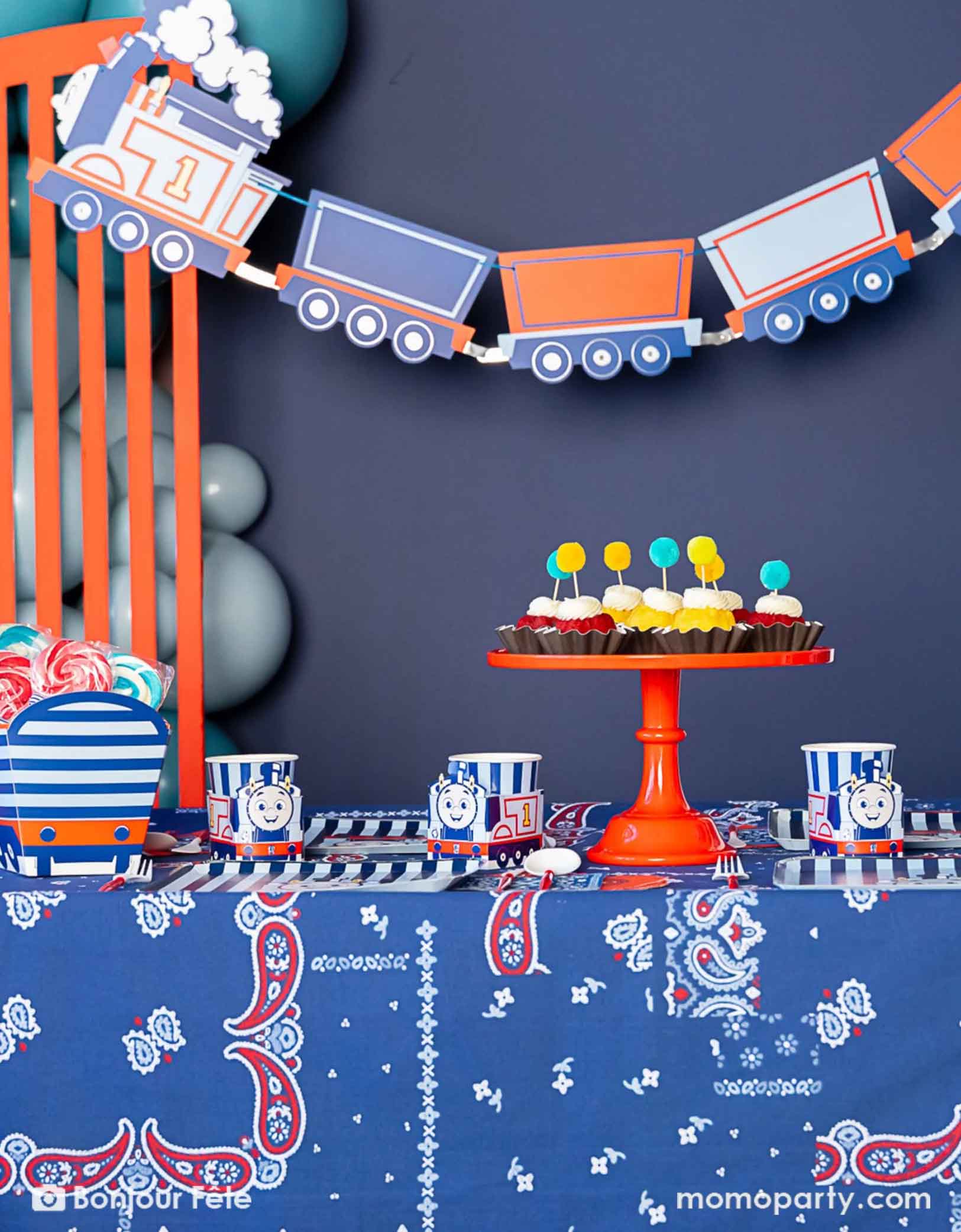 An adorable and festive Thomas the Train and Friends themed train party set up featuring Momopartys's Thomas the Train themed party supplies including party tableware of plates, cups, napkins treat boxes and party garland. The party table is covered with bandana inspired tablecloth to match the theme. In the middle of the table there's a cake stand holding cupcakes in yellow and red colors topped with pom pom toppers. In the back of is a blue balloon garland, making this a cute inspo for kid's train party.