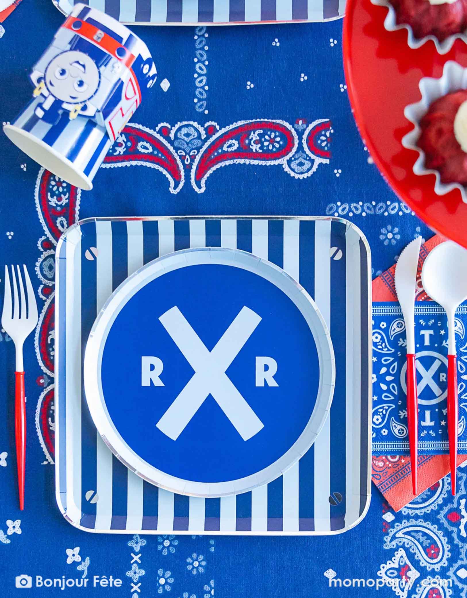 An overhead shot of an adorable and festive Thomas the Train and Friends themed train party table featuring Momopartys's Thomas the Train themed party  tableware including striped dinner plates, the crossroad round side plates, the 3D party cups, red cutlery set and bandana napkins. The party table is covered with bandana inspired tablecloth to match the theme, making this a perfect table setting inspo for a toddler's train themed birthday party.