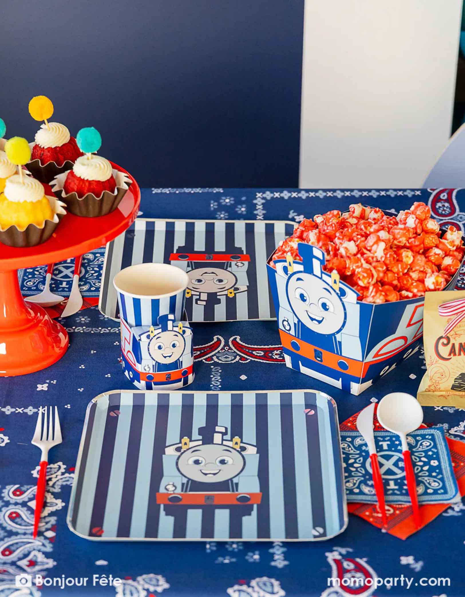 An adorable and festive Thomas the Train and Friends themed train party table featuring Momopartys's Thomas the Train themed party supplies including party tableware of striped plates, 3D cups, napkins and treat boxes. The party table is covered with bandana inspired tablecloth to match the theme. In the middle of the table there's a red cake stand holding cupcakes topped with pom pom toppers. In the back is a blue & red balloon garland, making this a perfect inspo for toddler's train themed birthday party.