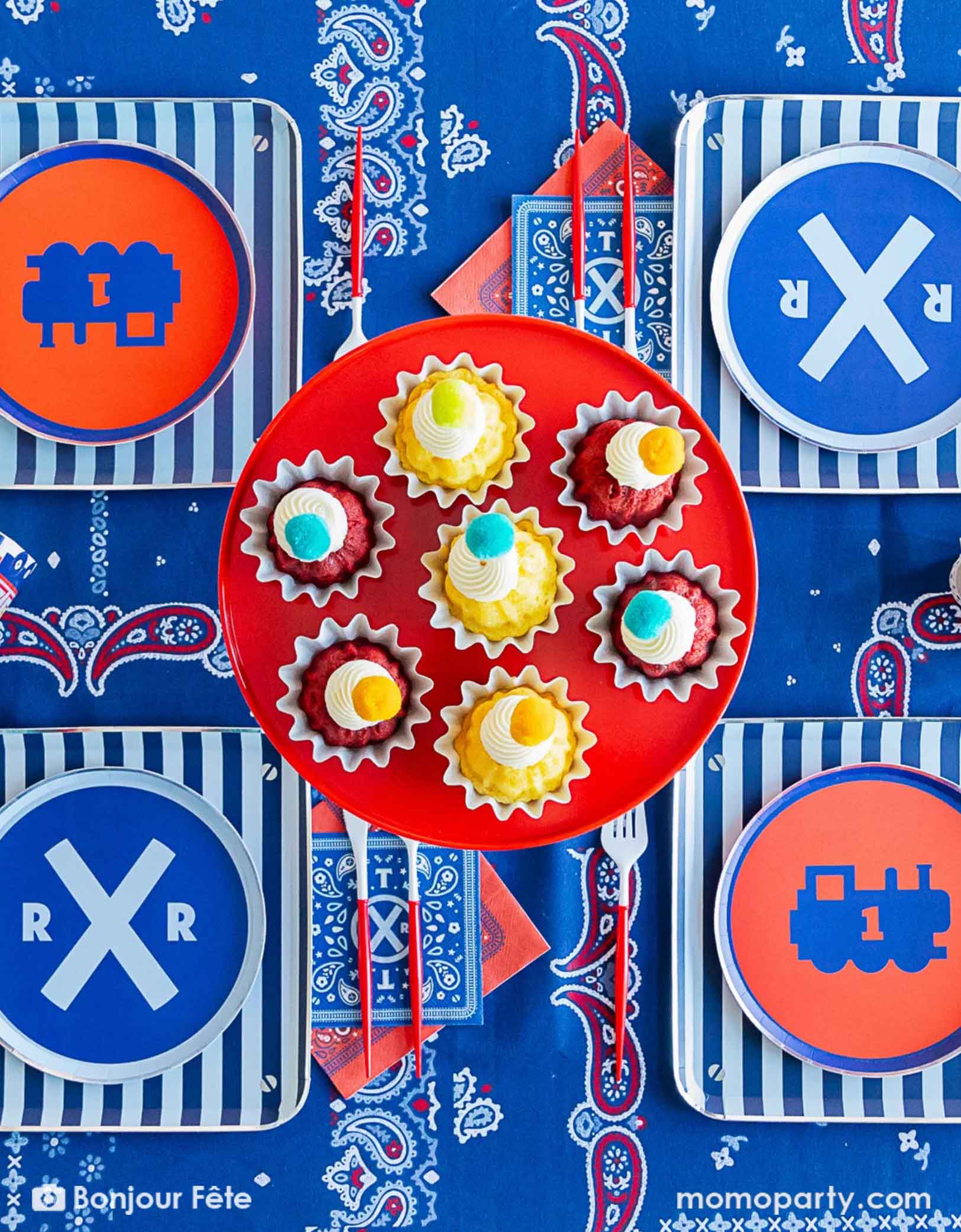 An overhead shot of an adorable and festive Thomas the Train and Friends themed train party table featuring Momopartys's train themed party tablewares including striped dinner plates, the crossroad round side plates, the 3D party cups, red cutlery set and bandana napkins. In the middle of the table there's a red cake stand holding cupcakes with pompoms. The table is covered with bandana inspired tablecloth, making this a perfect table setting inspo for a kid's train themed birthday party.