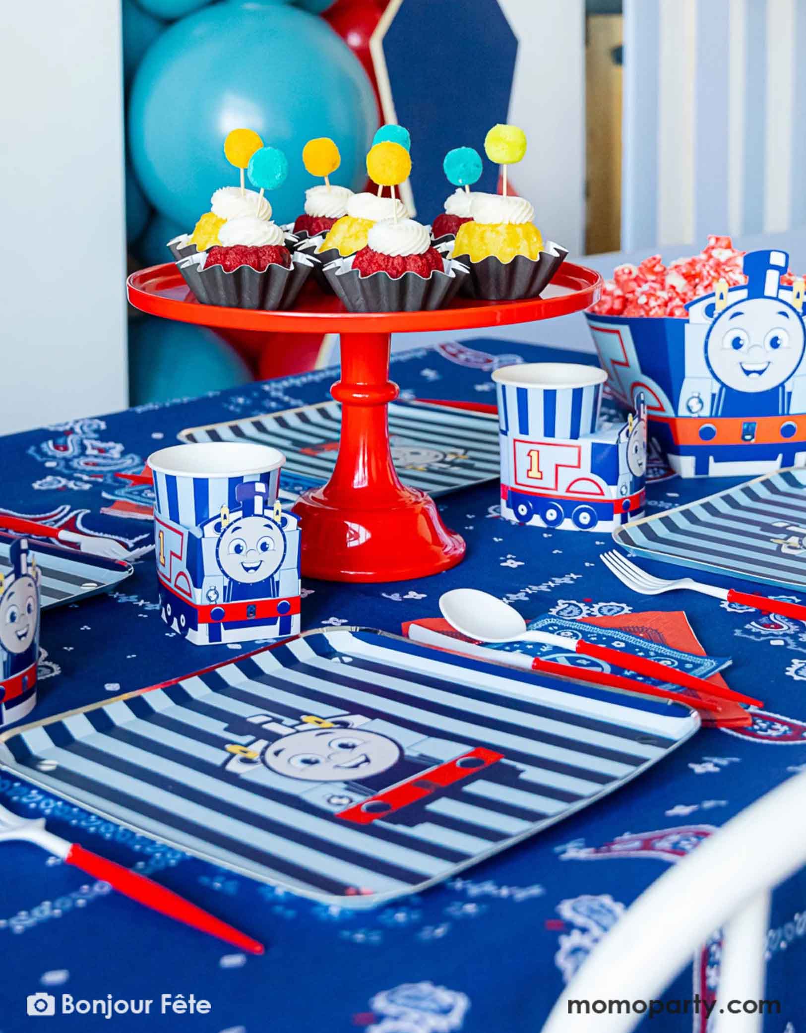 An adorable and festive Thomas the Train and Friends themed train party table featuring Momopartys's Thomas the Train themed party supplies including party tableware of plates, 3D cups, napkins treat boxes. The party table is covered with bandana inspired tablecloth to match the theme. In the middle of the table there's a red cake stand holding cupcakes topped with pom pom toppers. In the back is a blue & red balloon garland, making this a perfect inspo for toddler's train themed birthday party.