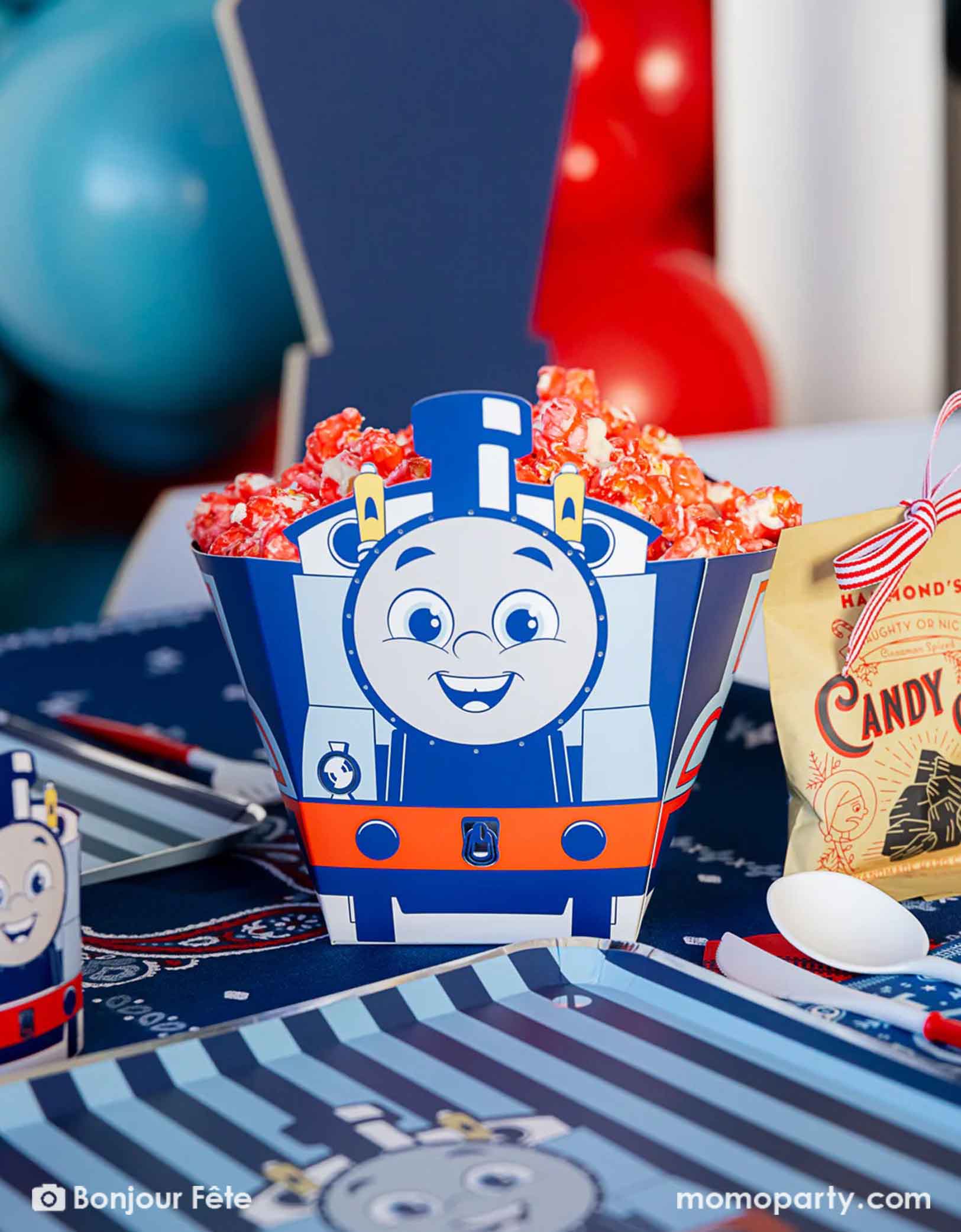 A close-up of the shot of Thomas the Train and Friends shaped treat box filled with red popcorn in a festive Thomas the Train and Friends themed train party. On the table there are various Thomas the Train themed party supplies from Momopartys including party tableware of plates, 3D cups, napkins treat boxes. The party table is covered with bandana inspired tablecloth to match the theme. In the back is a blue & red balloon cloud, making this a perfect inspo for a toddler's train birthday celebration.