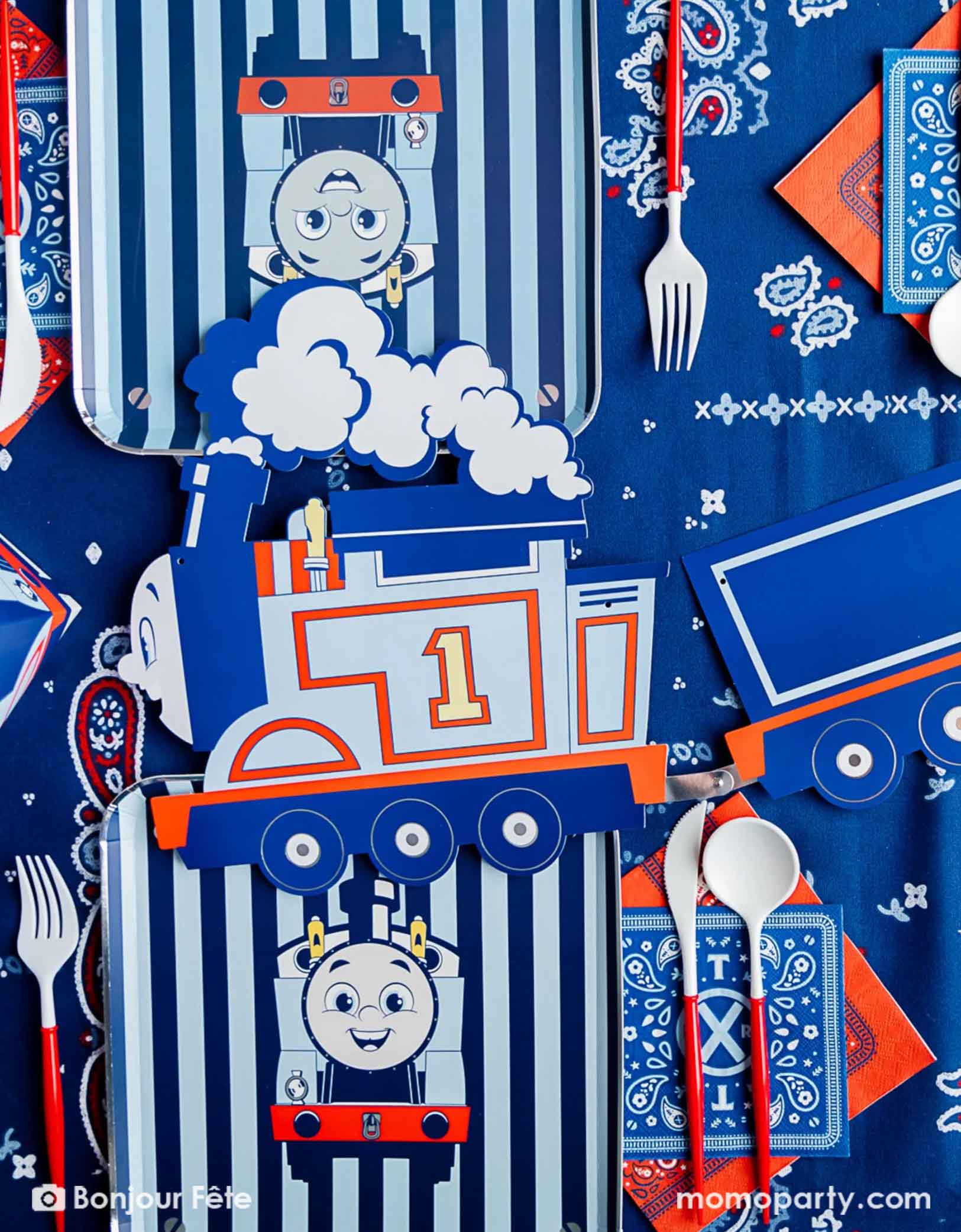An overhead shot of an adorable and festive Thomas the Train and Friends themed train party table featuring Momopartys's Thomas the Train themed party  supplies including striped dinner plates, red cutlery set, bandana napkins and a party garland in the middle. The party table is covered with bandana inspired tablecloth to match the theme, making this a perfect table setting inspo for a toddler's train themed birthday party.