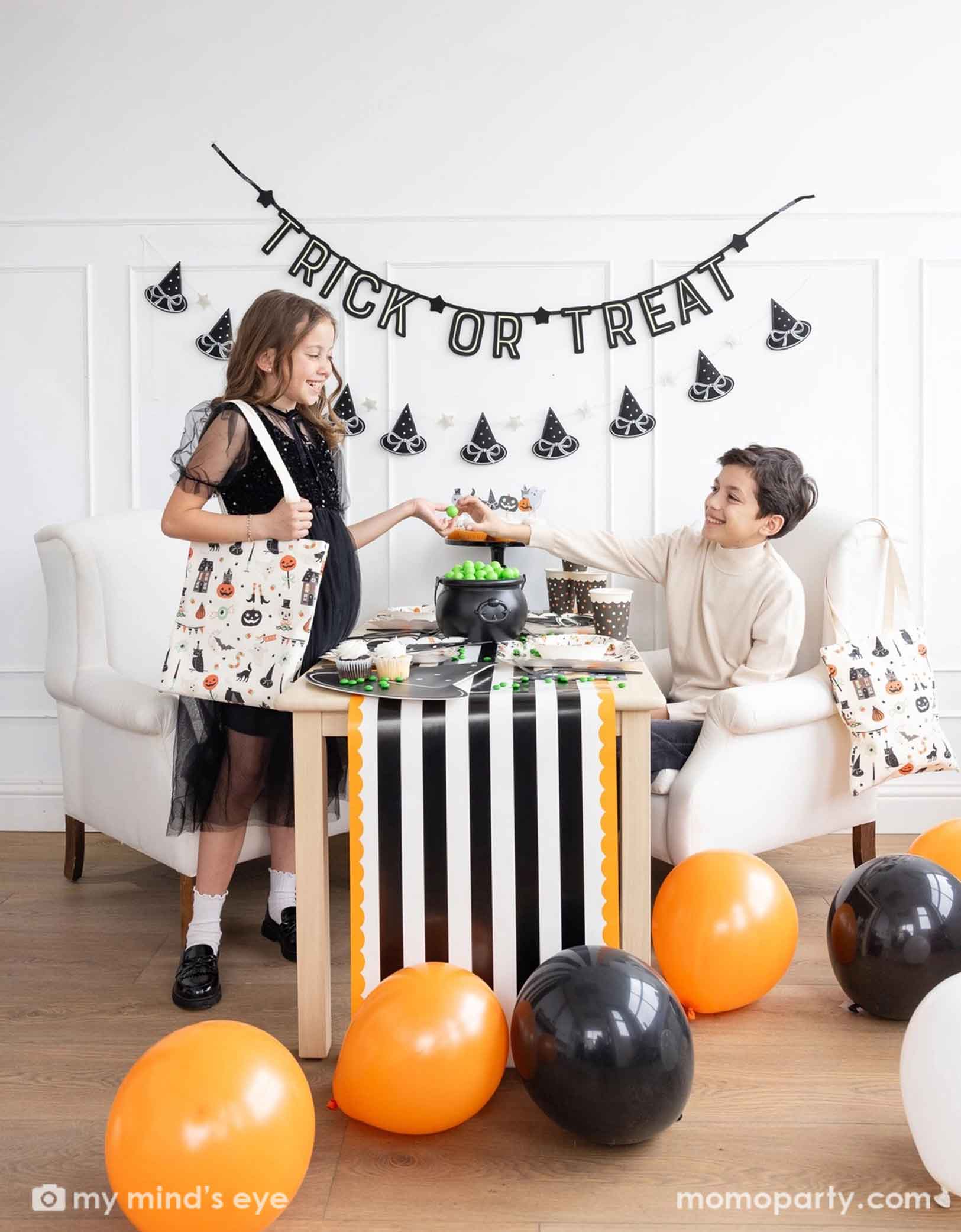 Two children at a Halloween-themed party with Momopartys's 'Trick or Treat' banner and the witch's hat garland hung on the white wall. There's a table decorated with multiple Halloween party supplies by Momopartys including witch's hat shaped bamboo tray, party cups and orange, black and white striped table runner draping from the table which some orange and black balloons next to. A cute and simple set up for kids to celebration Halloween at home.