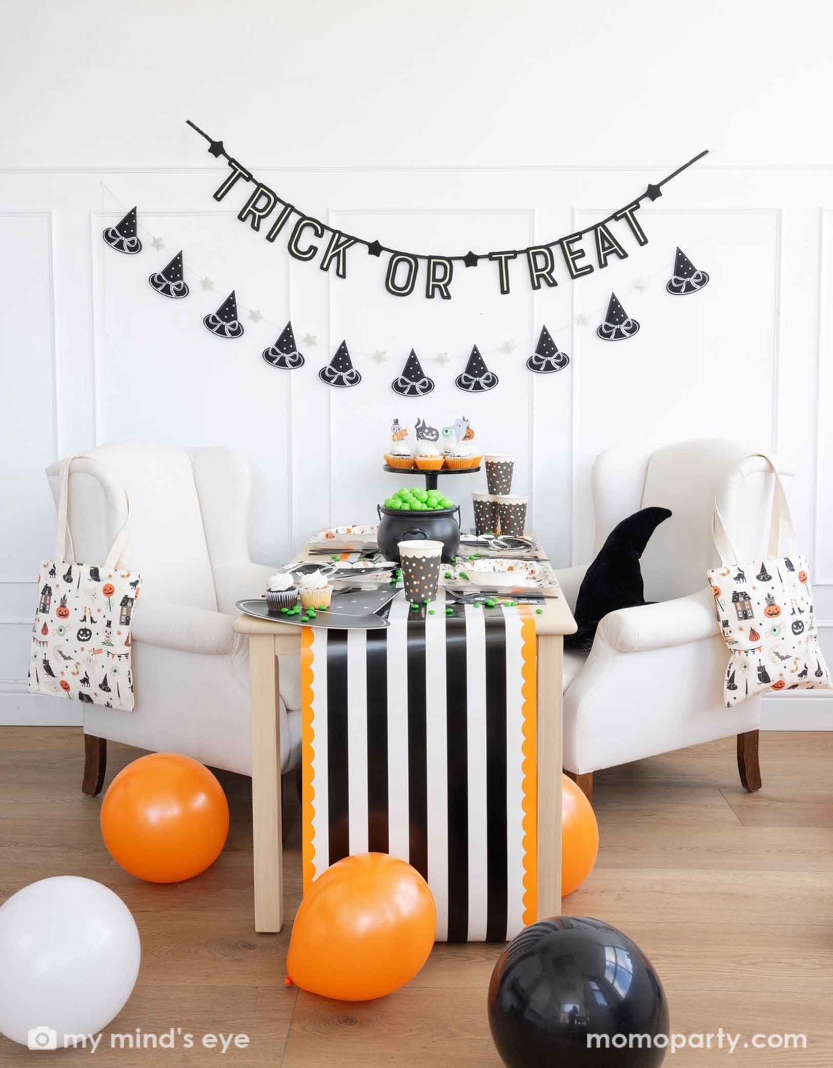 A Halloween-themed party with Momopartys's 6 ft 'Trick or Treat' glow in the dark banner and the witch's hat garland hung on the white wall. There's a table decorated with multiple Halloween party supplies by Momopartys including witch's hat shaped bamboo tray, party cups and orange, black and white striped table runner draping from the table which some orange and black balloons next to. A cute and simple set up for kids to celebration Halloween at home.