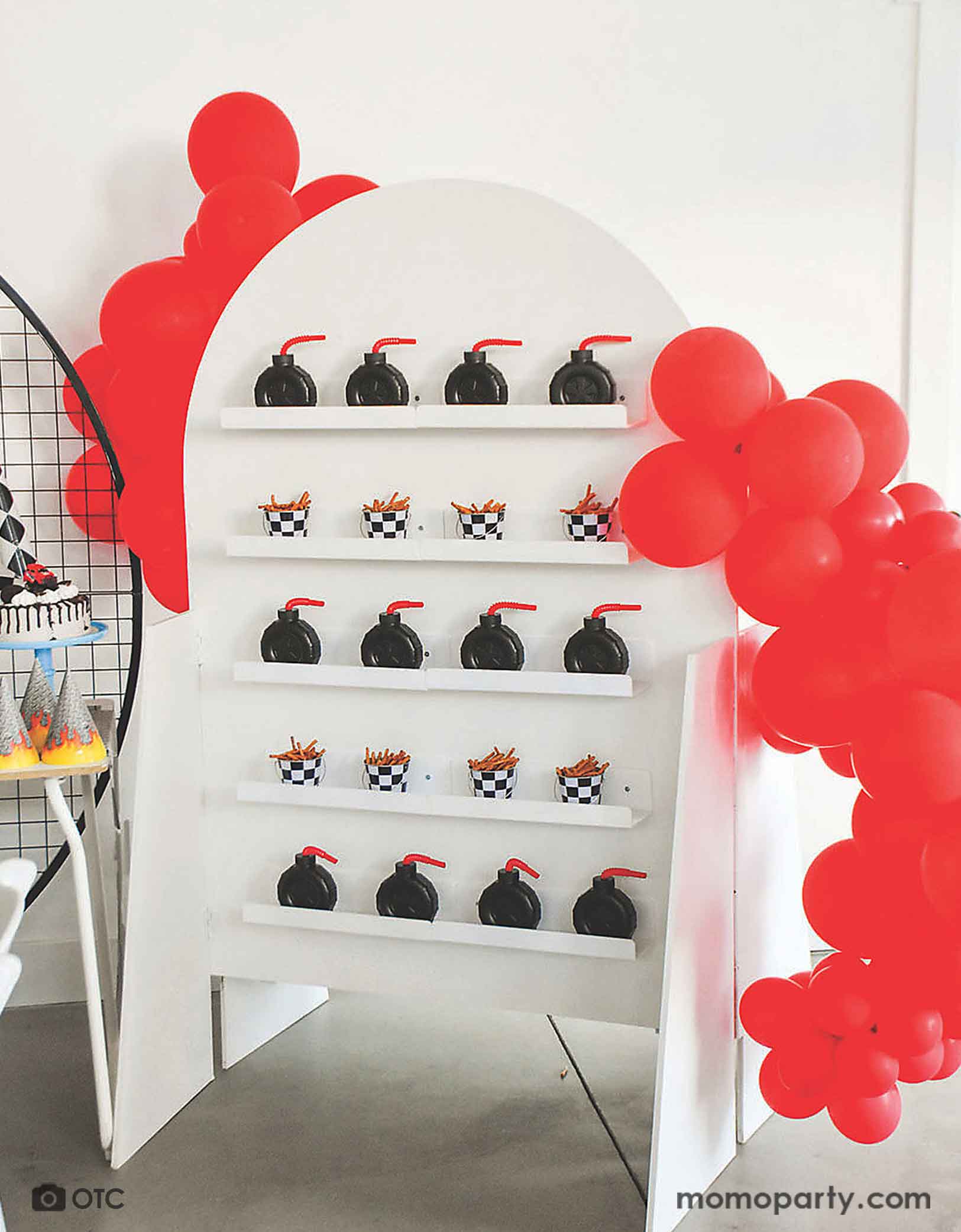 A kid's monster truck themed birthday party favor wall filled with Momopartys's 12 oz 4" tire truck shaped cups and mini checkered board buckets with pretzels. Around the wall, there are two red balloon garlands, making this a modern and clean set up for a fun kid's monster truck car themed birthday celebration!