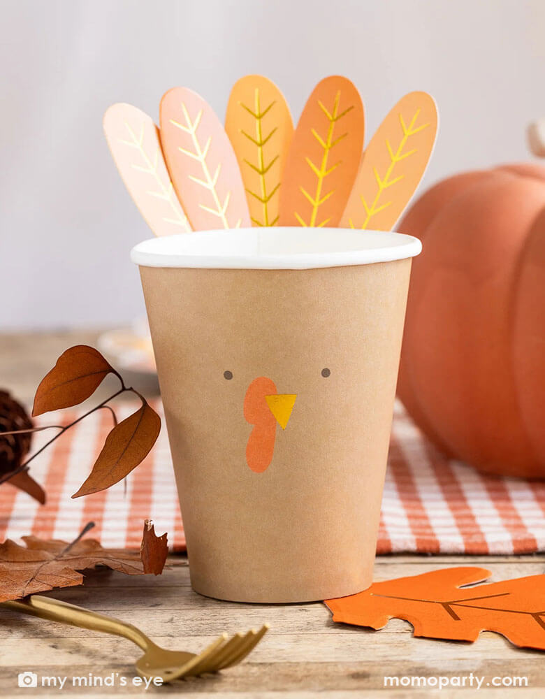 A Thanksgiving party table shot featuring Momopartys's 8 oz Harvest Turkey Paper Party Cup by My Mind's Eye. On the table there are some fall leaves decoration, an oak leaf shaped napkin, a pumpkin and an orange gingham checkered table runner, this cozy table setting makes a great inspiration for this harvest season. 