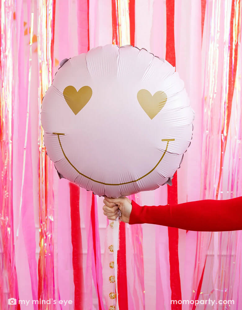 A hand in red sweatshirt holding Momopartys's heart eyes foil balloon by My Mind's Eye, in front of a wall decorated with red, pink and iridescent streamers for a fun Valentine's Day celebration.