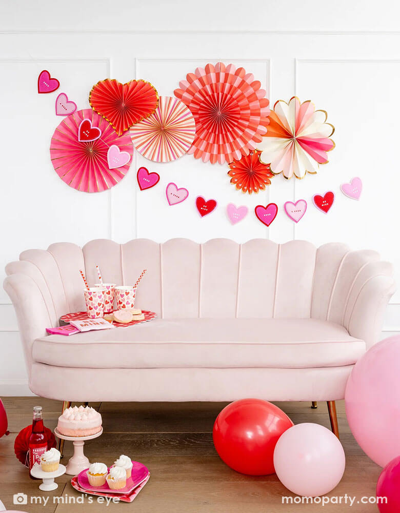 A gorgeous Valentine's party celebration featuring heart you paper fans on the wall with a heart shaped banner. Momopartys's Valentine scatter party cups and straws and napkins are on a pink sofa, in the front there is a small pink cake and some cupcakes with pink soda drinks. Around the sofa the room is decorated with pink and red latex balloons, ready for a celebration on a sweet Valentine's Day.