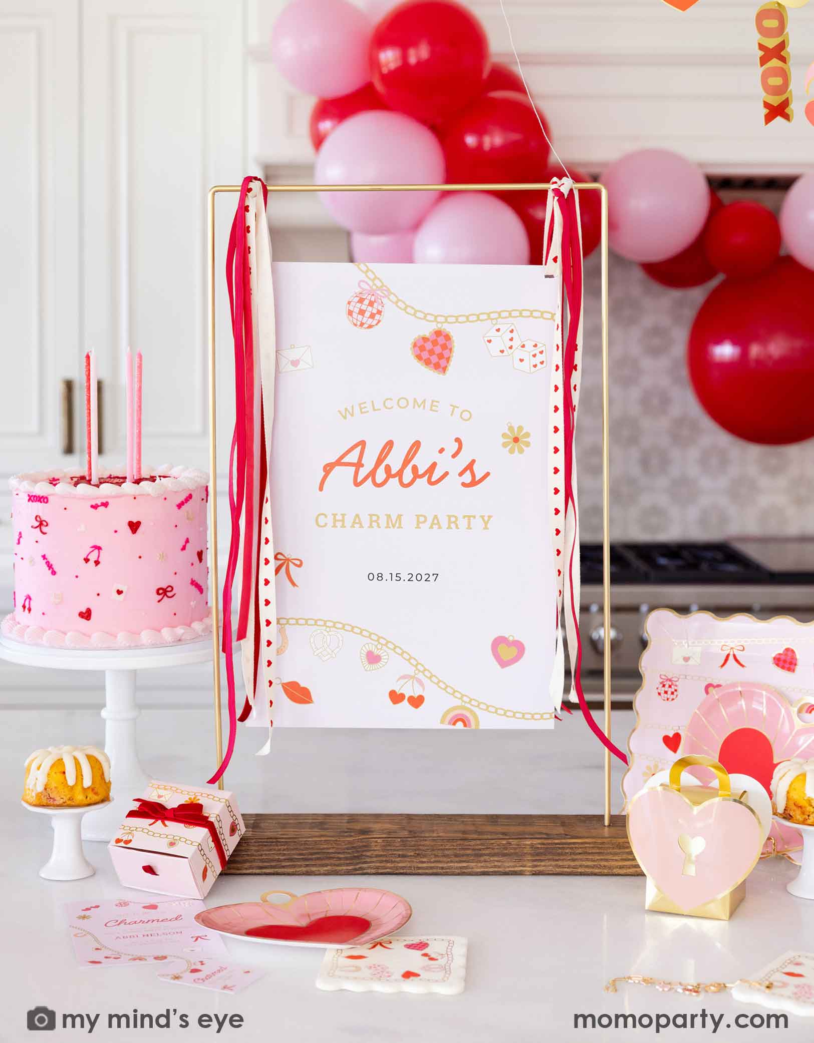 A Valentine's Day celebration decorative setup with a pink cake, red and pink balloons, and 'Welcome to Abbi's Charm Party' sign next to various Valentine's Day party supplies from Momopartys including the heart charm shaped plates, matching napkins and treat bags. Making this a festive scene for kid's Valentine's Day party or a Galentine's Day celebration.