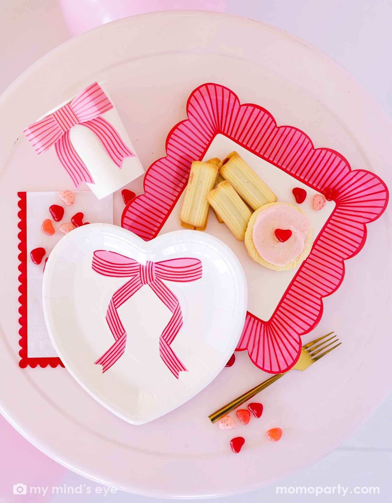 A overhead shot of Momopartys's bow heart Valentine's Day party collection including light pink heart shaped plate, bow party cup, striped scallop edged plate and scallop napkins. Around the tableware there are some heart shaped candies, cookies and some Valentine's Day themed snacks.