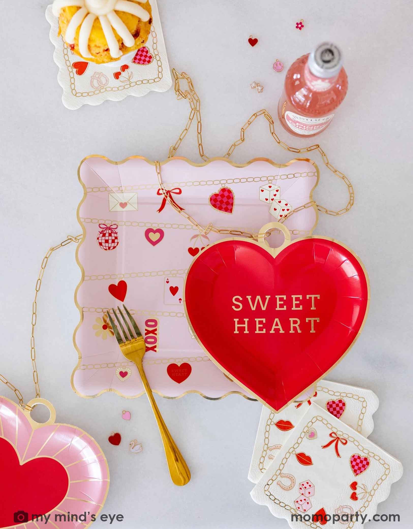 An overhead shot of a Valentine's Day party tablescape featuring various Valentine's Day party supplies from Momopartys including the red heart shaped plate with 'Sweet Heart' text and Valentines Charm Scalloped Square Plate and Valentines Charm Small Napkins by My Mind's Eye, surrounded by decorative Valentine's items, charms, snacks and drinks on a light background.