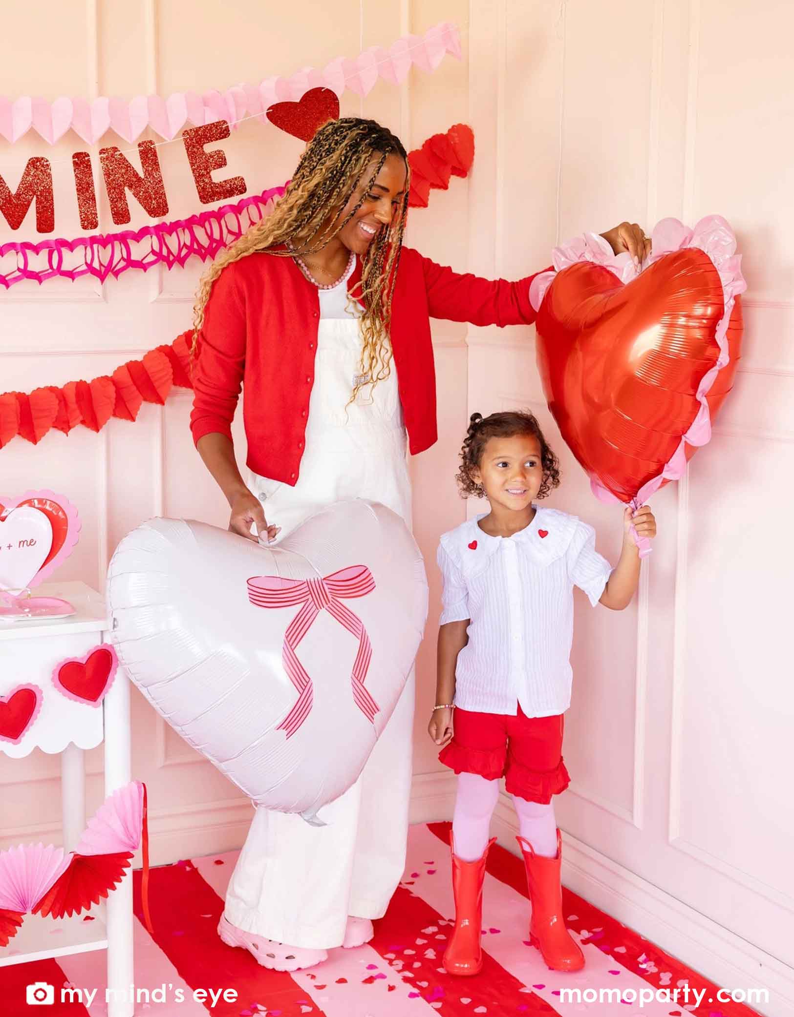A mom and her child in red and pink outfits holding Momopartys's 22" heart-shaped balloons by My Mind's Eye. With 'BE MINE' party banner and red and pink tissue paper garlands in the back on a pink wall. In the front there's a dessert table filled with Valentine's Day party supplies and heart shaped decorations. A perfect inspiration for a sweet Valentine's Day celebration with family and kids.
