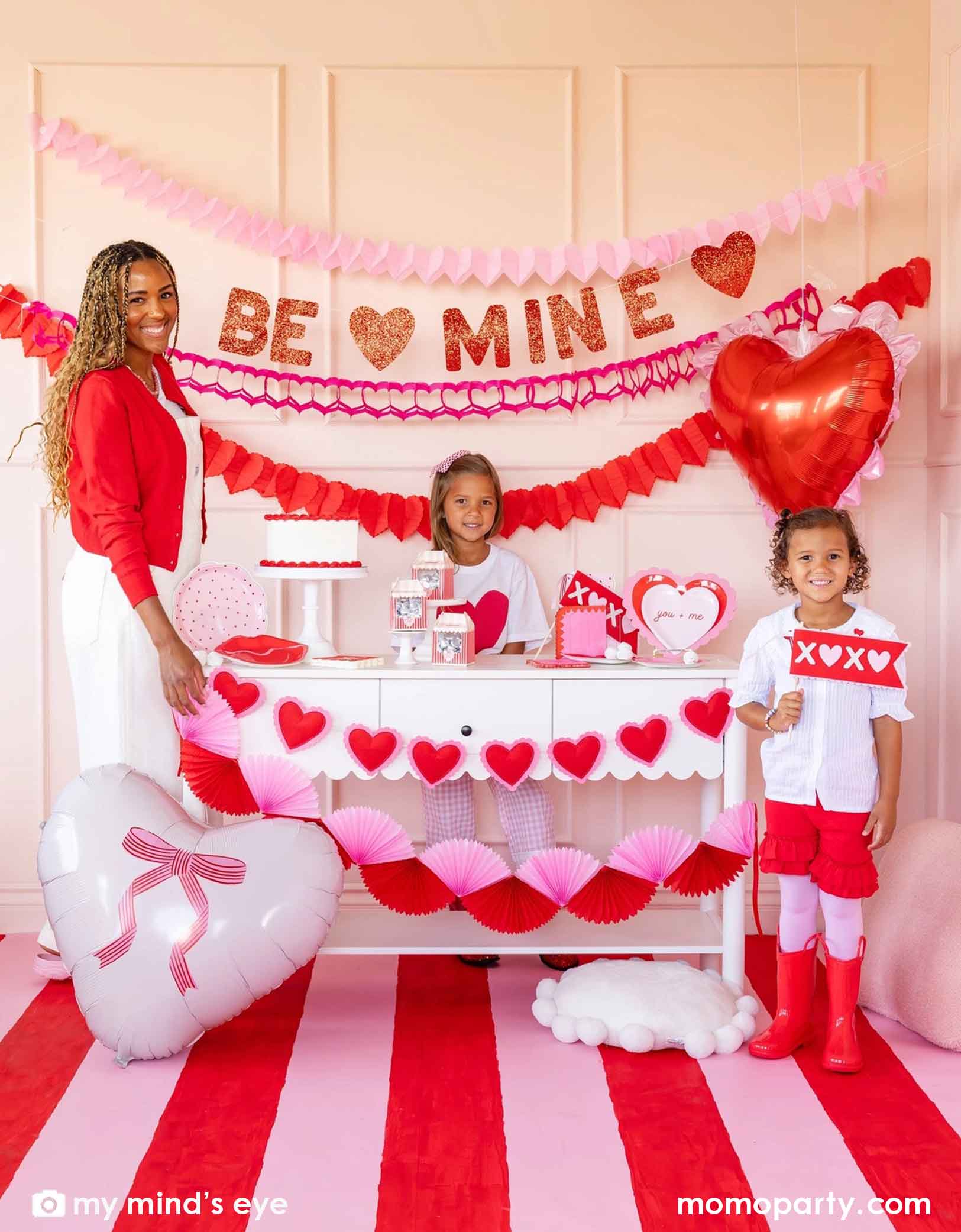 A mom and her kids at a Valentine's Day party set up featuring various Momopartys's Valentine's Day party supplies and decorations including 22" heart-shaped balloons by My Mind's Eye, 'BE MINE' party banner, red and pink tissue paper garlands in the back on a pink wall. In the front there's a dessert table filled with Valentine's Day party supplies and heart shaped banner decorations in red and pink. A perfect inspiration for a sweet Valentine's Day celebration with family and kids.