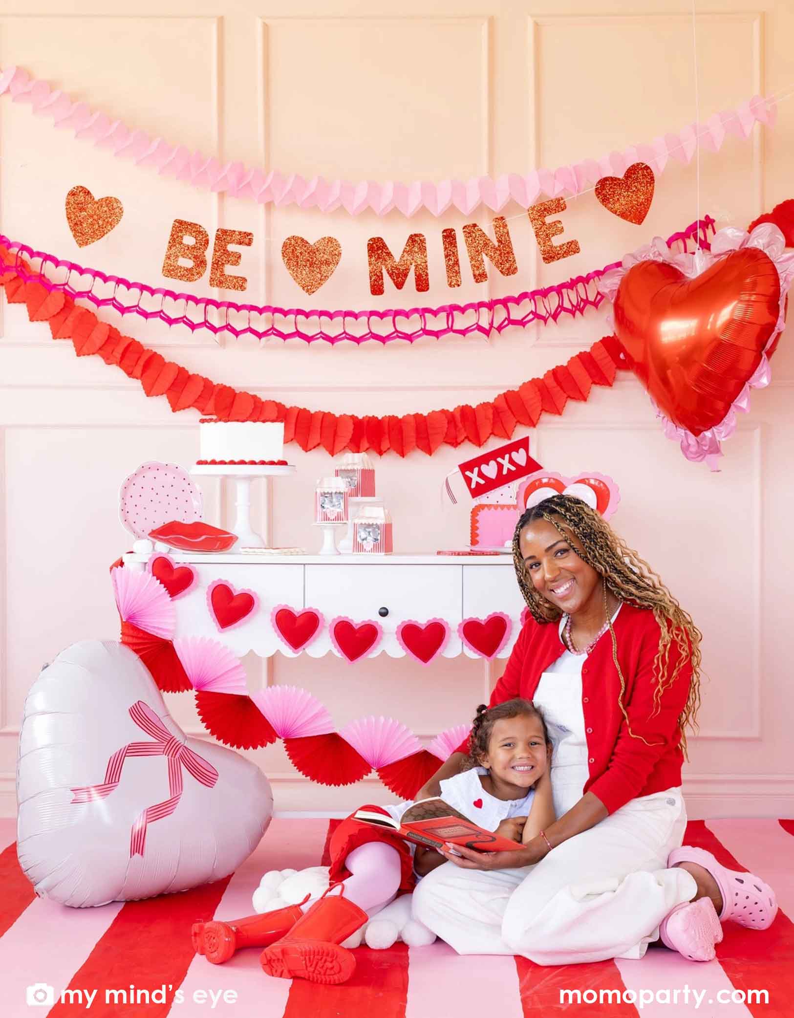 A mom and her kid sitting on a red and white striped rug at a Valentine's Day party set up featuring various Momopartys's Valentine's Day party supplies and decorations including 22" heart-shaped balloons by My Mind's Eye, 'BE MINE' party banner, red and pink tissue paper garlands in the back on a pink wall. In the front there's a dessert table filled with Valentine's Day party supplies and heart shaped banner decorations. A perfect inspiration for a sweet Valentine's Day celebration with family and kids.