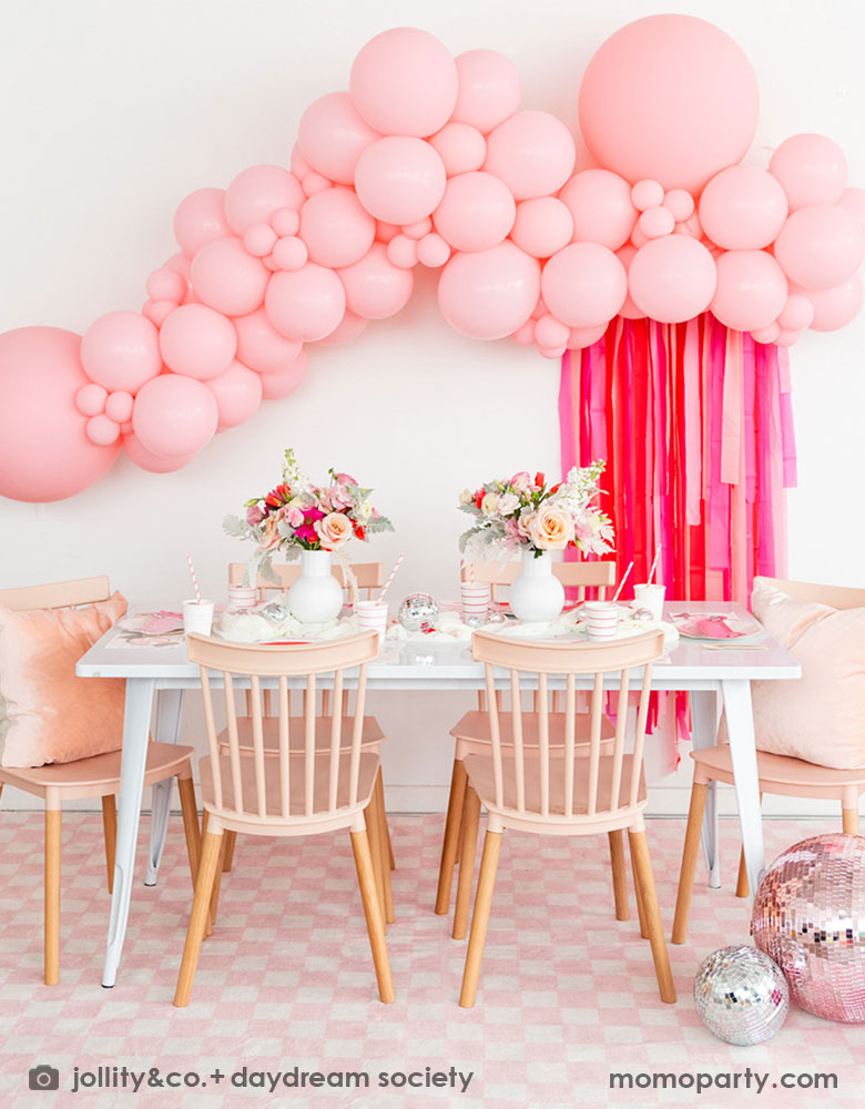 A beautiful Valentine's Day party set up featuring a gorgeous pink balloon garland adorned with rose pink streamer. On the party table there are Momopartys's red striped tableware including cups, plates, napkins, along with some pretty pink and red flower arrangement. Along with light pink wooden chairs with pink pillows, pink checkered rug and large pink disco balls, this makes a great inpo for a darling Valentine's Day celebration for girl's gatherings. 
