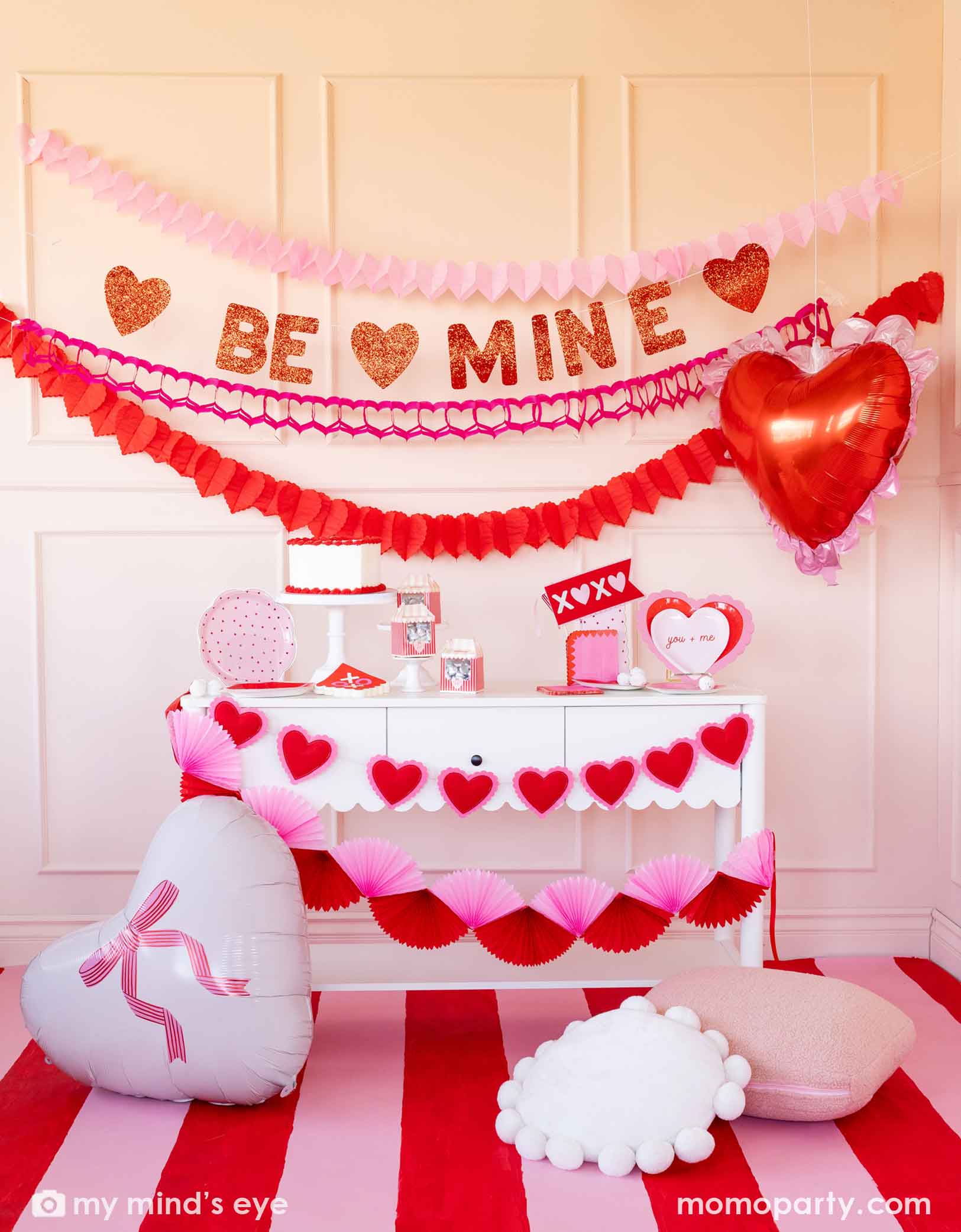 A festive Valentine's Day party set up featuring various Momopartys's Valentine's Day party supplies and decorations including two 22" heart-shaped balloons by My Mind's Eye, 'BE MINE' party banner, red and pink tissue paper garlands in the back on a pink wall. In the front there's a dessert table filled with Valentine's Day party supplies and heart shaped banner decorations. A perfect inspiration for a sweet Valentine's Day celebration with family and kids.