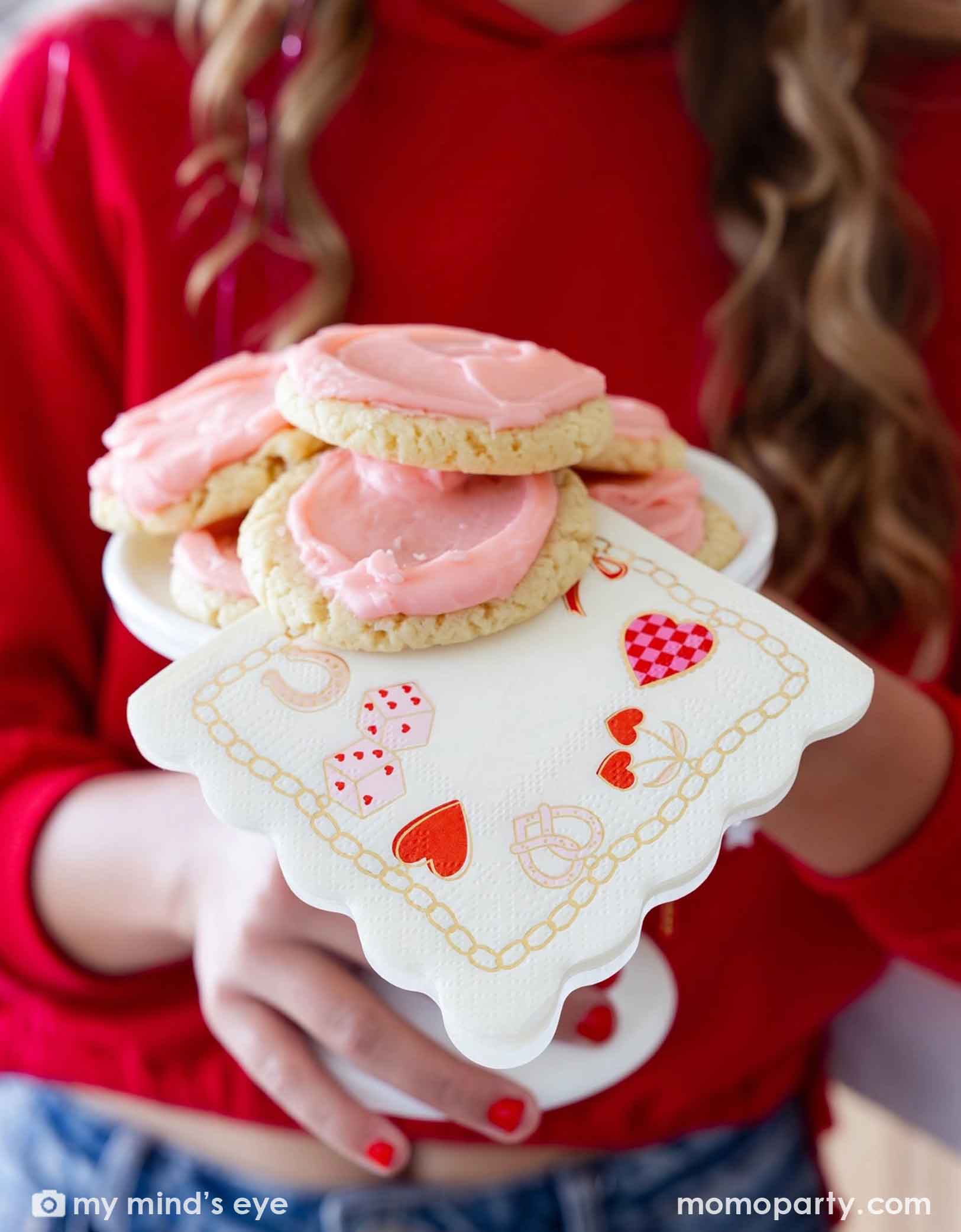 A girl in red sweat shirt with red nail polish  holding a small cake stand with pink frosted cookies shaped like hearts and Momopartys's 5" x 5" Valentines Charm Small Napkin featuring various Valentine's Day themed illustrations at a Valentine's Day or Galentine's Day celebration party.