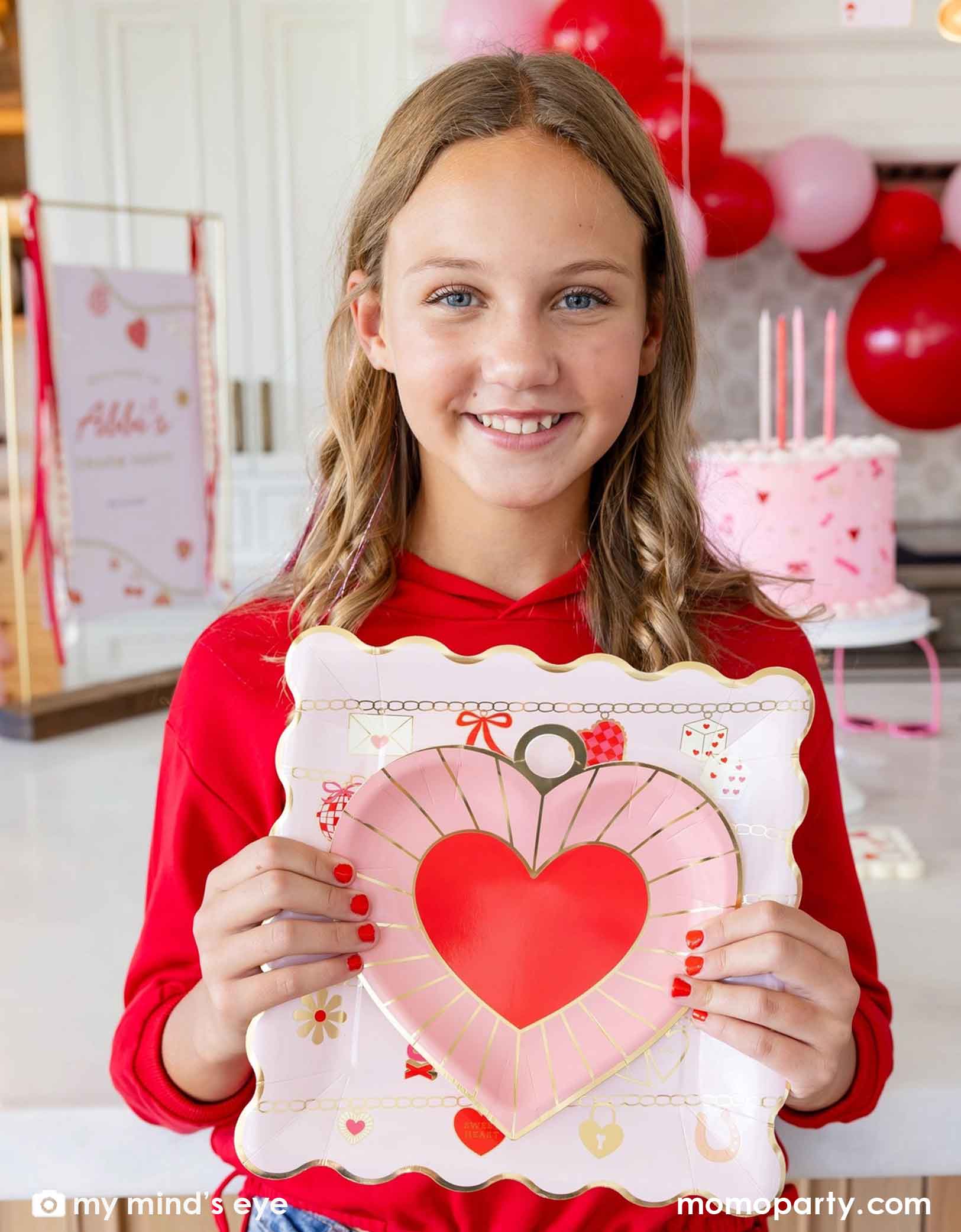 A young blonde girl in a Valentine's Day party scene holding Momopartys's pink heart-shaped plate and pink square plate with Valentine's Day balloon garland and a pink buttercream cake in the background.