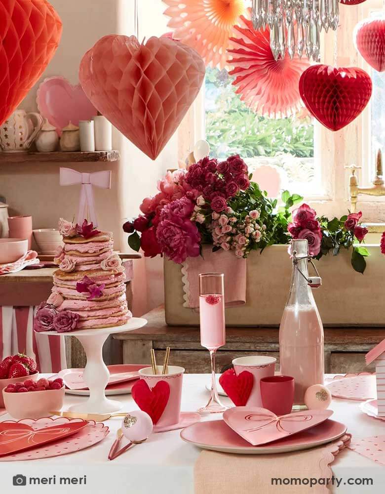 A festive Valentine's Day party featuring a table filled with Momopartys's red and pink heart shaped party supplies including the scallop edged dinner plates, napkins, heart with bow plates and heart honeycomb party cups, in the middle is a pink pancake tower adorned with pink and red flowers topped with a pink bow.  Above the table, there are heart shaped honeycombs and paper fans hanging from the ceiling next to a big window, making this a great inspo for Valentine's Day celebration.