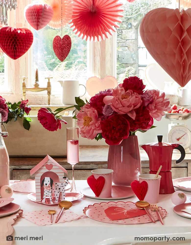 A festive Valentine's Day party featuring A table filled with Momopartys's red and pink heart shaped party supplies including the scallop edged dinner plates, napkins, and heart honeycomb party cups, in the middle is a vase filled with red and pink roses. above the table, there are heart shaped honeycombs and paper fans hanging from the ceiling next to a big window, making this a great inspo for Valentine's Day celebration.