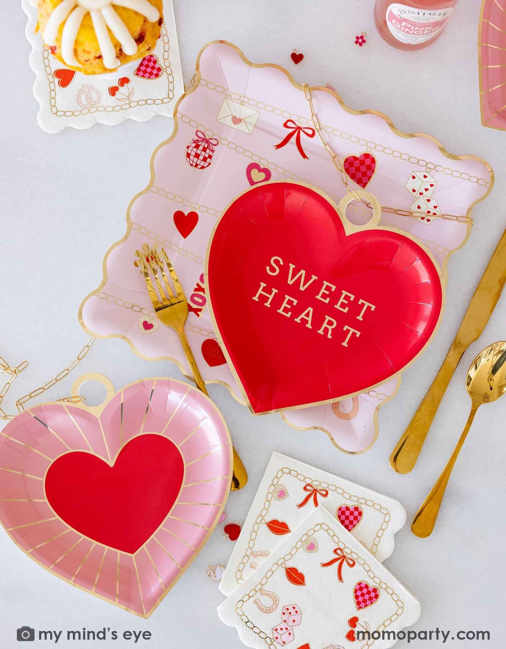 A closeup of an overhead shot of a Valentine's Day party tablescape featuring various Valentine's Day party supplies from Momopartys including the red heart shaped plate with 'Sweet Heart' text, pink heart shape plates, Valentines Charm Scalloped Square Plate and Valentines Charm Small Napkins by My Mind's Eye, surrounded by decorative Valentine's items, charms, snacks, drinks and gold utensils on a light background.