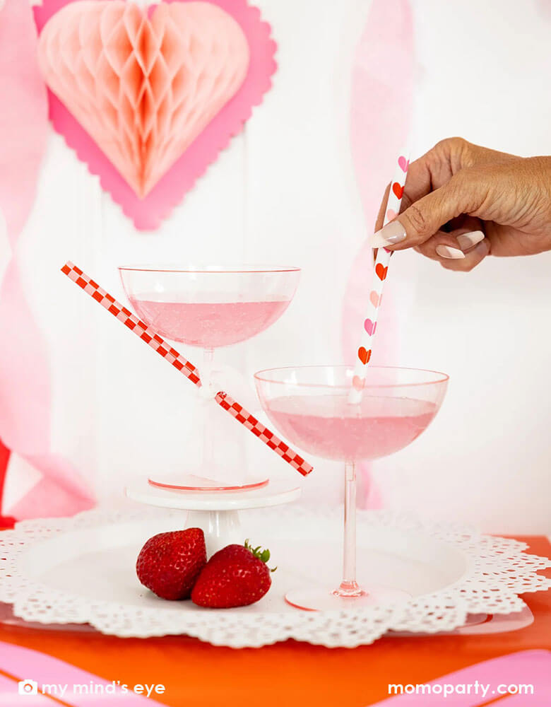 Two cocktail glasses with pink drink for a Valentine's Day celebration. A lady is mixing and stirring the drink with Momopartys's heart you reusable straws by My Mind's Eye. In the back there's honeycomb heart shaped hanging decoration, making it a gorgeous inspiration for a sweet Valentine's Day party.