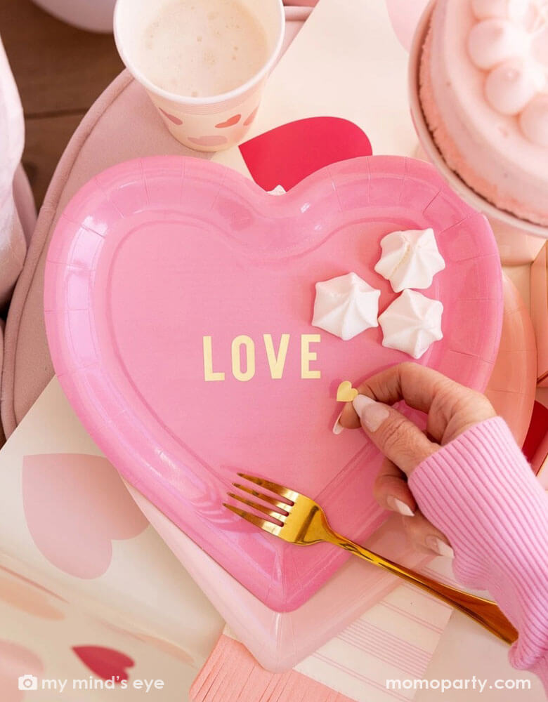 A Valentine's Day celebration party featuring a pink heart shaped plate with "LOVE" on it, layered on Momopartys's Scratch Off pink heart table runner. A girl's hand in a pink sweater reaches to get some meringue desserts. On the table you can see some darling pink heart themed tableware including party cups, napkins and plates, making this a adorable tablesetting inspo for a darling Valentine's Day celebration.