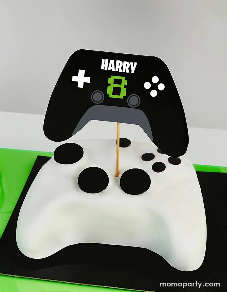 A video game console controller shaped fondant cake topped with Momopartys's Video Game Console Controller shaped personalized Cake Topper by Hooty Balloo. Come with two sticker sheets, you can customize this cake topper with the birthday person's name and age. In a classic black and green colors, it's perfect for kid's video game themed birthday party.