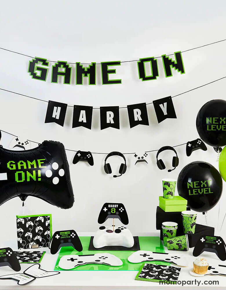 A kid's video game themed 8th birthday party set up featuring Momopartys's video game themed party supplies including Game On foil balloon, Next Level black latex balloons, personalized game on party banner on the wall. On the table there are white game controller shaped plates, along with black and green game controller motif party cups and napkins, in the middle a controller shaped birthday cake is topped with controller shaped personalized cake topper.