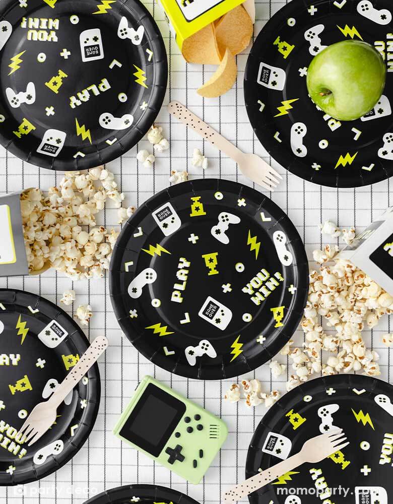 A video game themed party table features Momopartys's 9" Video Game Large Paper Plates by Party Deco. In the classic black color these round paper plates features gaming devices and controller design pattern, with pixelated words like "YOU WIN!" AND "PLAY" with neon lightning bolt symbols. On the checkered grid table, there are gaming snacks like popcorn and chips and some green apples along side some speckled wooden forks, a modern and stylish party table look for kid's video game themed birthday party.