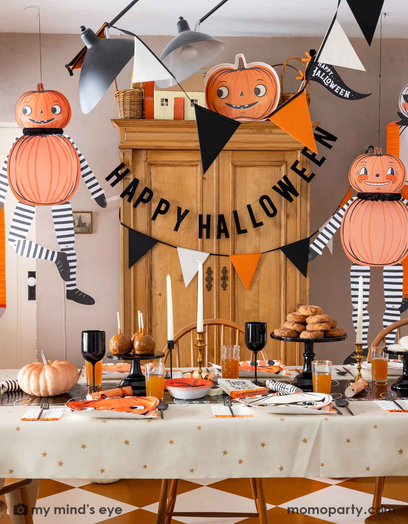 A festive Halloween-themed dining room set up with various Halloween decorations and party tablewares from Momopartys including jumbo Halloween paper hanging characters, orange, black and white felt banner and Happy Halloween banner hung  above. On the table there are vintage halloween party plates, napkins, party crackers. With donuts on the cake stand, black wine glasses, it creates a spooky fun vibe for a Halloween celebration for both kids and adults. 