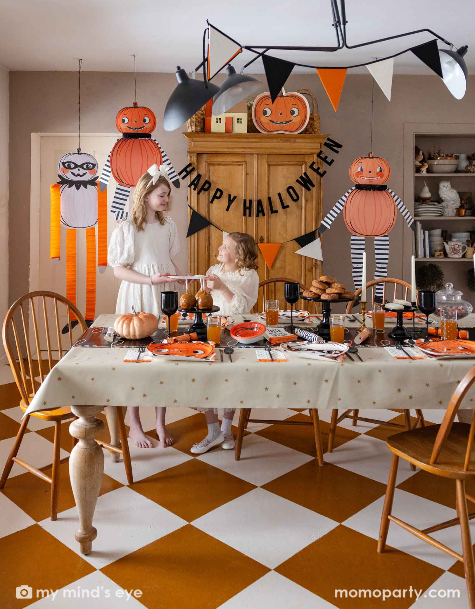 Two girls in a festive Halloween-themed dining room set up with various Halloween decorations and party tablewares from Momopartys including jumbo Halloween paper hanging characters, orange, black and white felt banner and Happy Halloween banner hung  above. On the table there are vintage halloween party plates, napkins, party crackers. With donuts on the cake stand, black wine glasses, it creates a spooky fun vibe for a Halloween celebration for both kids and adults. 