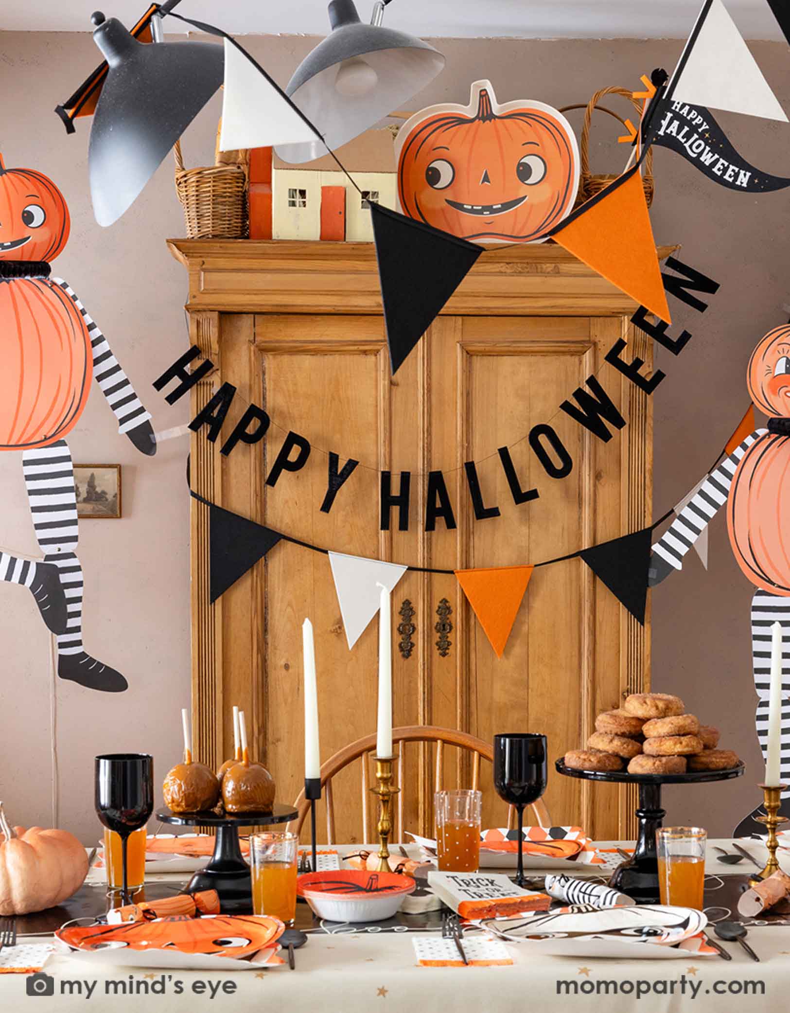 A festive Halloween-themed dining room set up with various Halloween decorations and party tablewares from Momopartys including jumbo Halloween paper hanging characters, orange, black and white felt banner and Happy Halloween banner hung  above. On the table there are vintage halloween party plates, napkins, party crackers. With donuts on the cake stand, black wine glasses, it creates a spooky fun vibe for a Halloween celebration for both kids and adults. 
