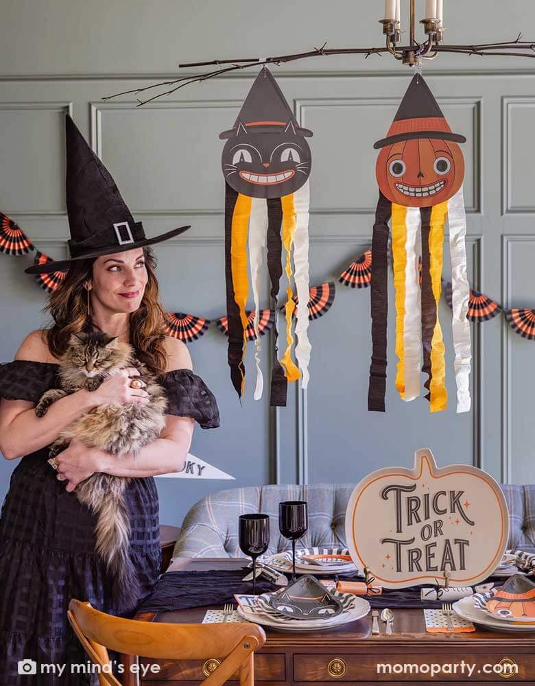 A lady dressed as a witch holding her cat in a Halloween party set up featuring vintage Halloween party supplies and decorations from Momopartys including the giant vintage Halloween character hanging decoration streamers and the black and orange bunting banner on the wall as the backdrop. On the table, there's a huge pumpkin shaped bamboo serving plate that says trick or treat on it. With vintage Halloween themed plates, napkins and glasses. This makes a great inspo for a fun Halloween gathering at home.