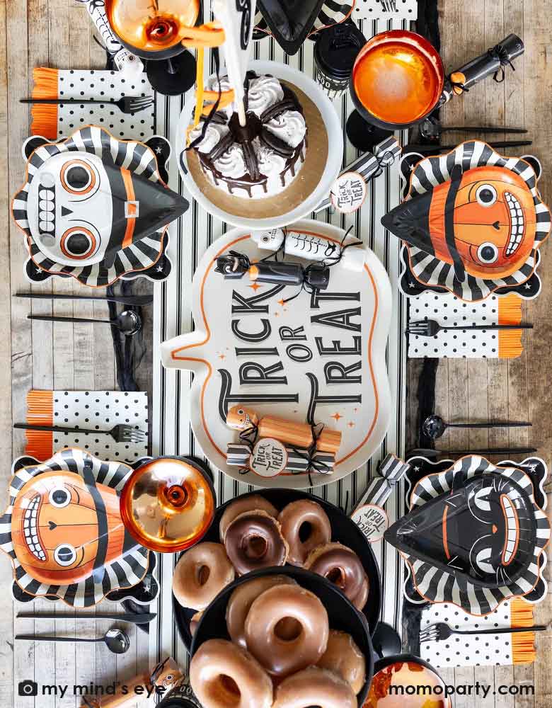 A overhead shot of a festive Halloween party table featuring Momopartys's vintage Halloween character shaped plates on the black starburst dinner plates and night sky scalloped plates, plus black dotted napkins with orange fringe. On the table there are some vintage Halloween party crackers on "Trick or Treat" pumpkin shaped bamboo serving tray on the black and white striped table runner. With delicious treats like cakes, donuts and apple cider, it's ready for a spooky fun Halloween celebration!