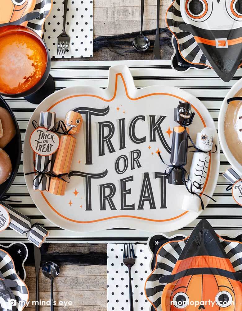A close up of a festive Halloween party table featuring Momopartys's vintage Halloween party supplies including Halloween party crackers in the designs of a pumpkin, a black at and a skeleton on  the"Trick or Treat" pumpkin shaped bamboo serving tray on the black and white striped table runner. Around the centerpiece were the Halloween character shaped plates layered with black starburst plates. With delicious treats like cakes, donuts and apple cider, it's ready for a spooky fun Halloween celebration!