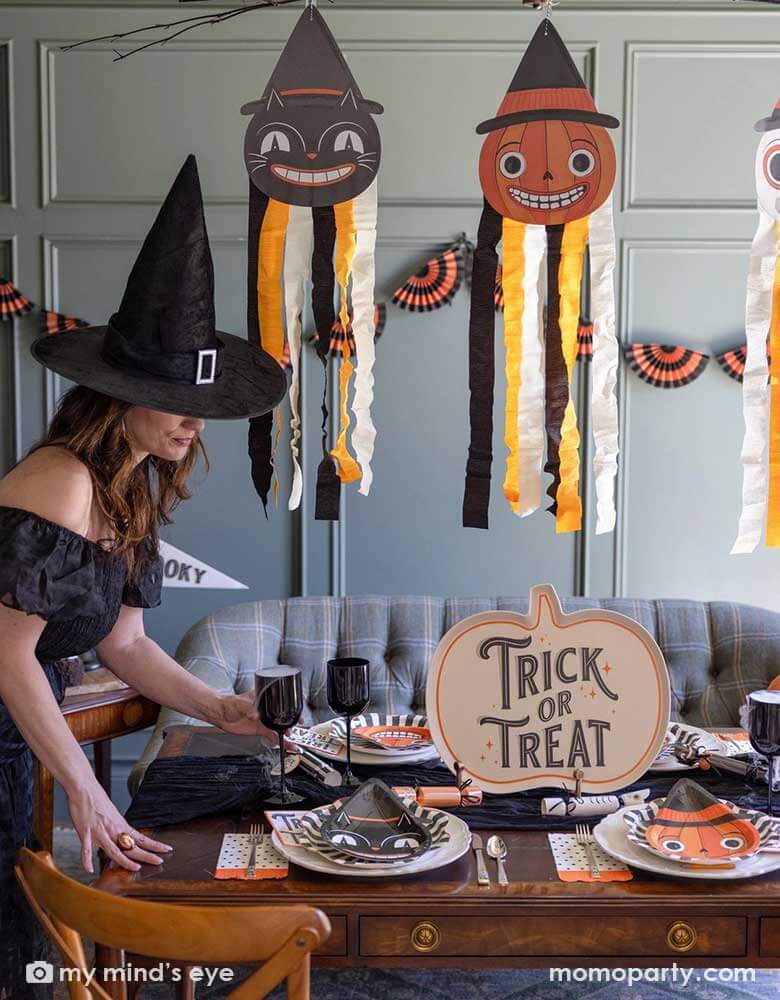 A lady dressed as witch setting up a Halloween party table in a vintage inspired Halloween party featuring Halloween party supplies and decorations from Momopartys including the giant vintage Halloween character hanging decoration streamers and the black and orange bunting banner on the wall as the backdrop. On the table, there's a huge pumpkin shaped bamboo serving plate that says trick or treat on it. With vintage Halloween themed plates, napkins and glasses.